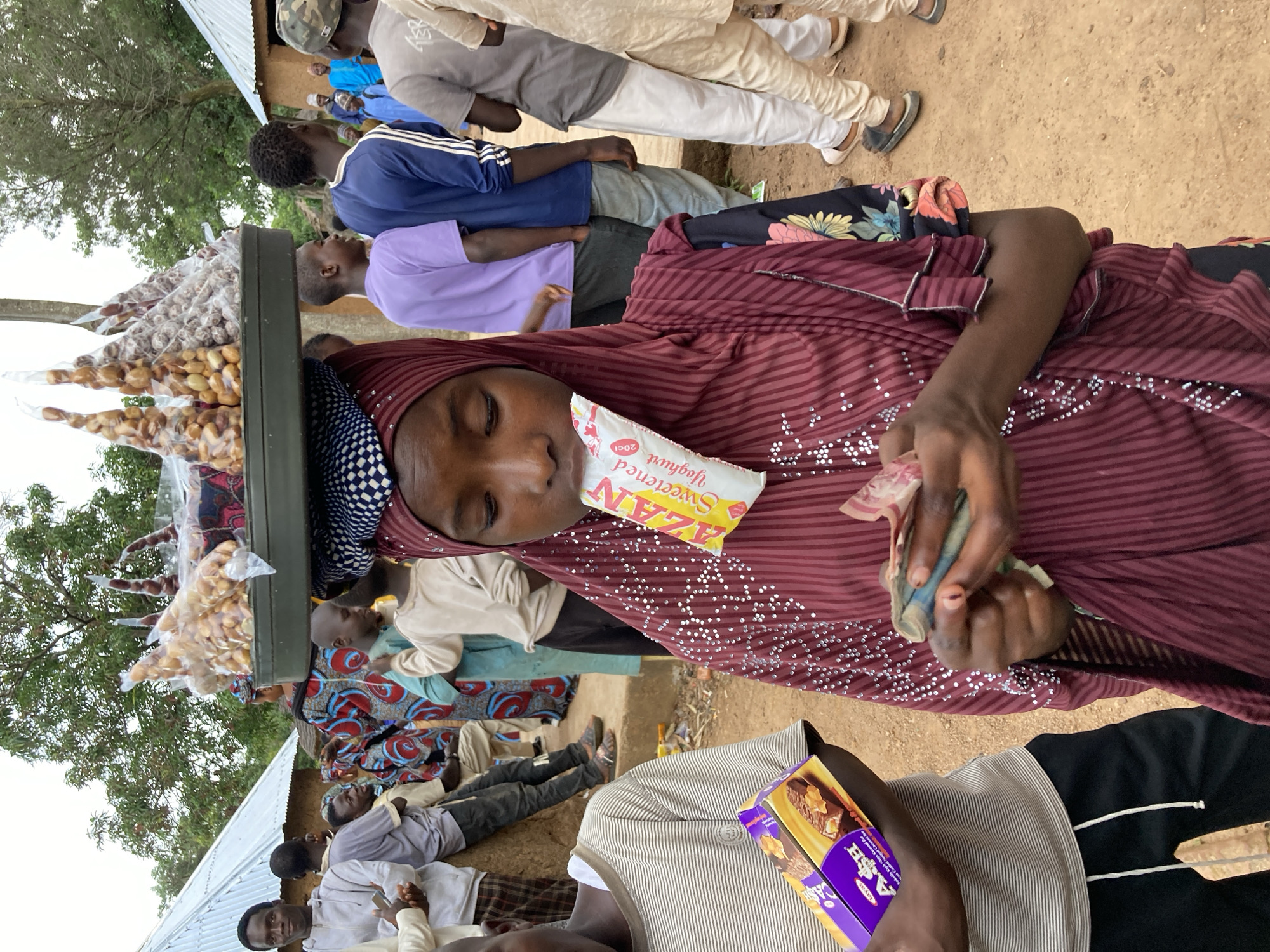 In an outdoor setting that appears to be a marketplace or a communal gathering space in Jos, Nigeria, a young girl wearing a maroon hijab and a patterned dress is the central subject. She is holding cash in her hands, seemingly engaged in a transaction, and has a packet of "AZAN Sweetened Yoghurt" in her mouth. A tray balanced on her head is laden with numerous plastic-wrapped packages of snacks, likely nuts. To her left, a boy in a striped shirt holds a purple box of what appears to be a chocolate bar. Around them, a diverse group of people are visible, some in casual attire and others in traditional clothing, suggesting a vibrant community scene. The background includes buildings with corrugated iron roofs and mud walls, characteristic of the local architecture. The ground is unpaved and dusty.