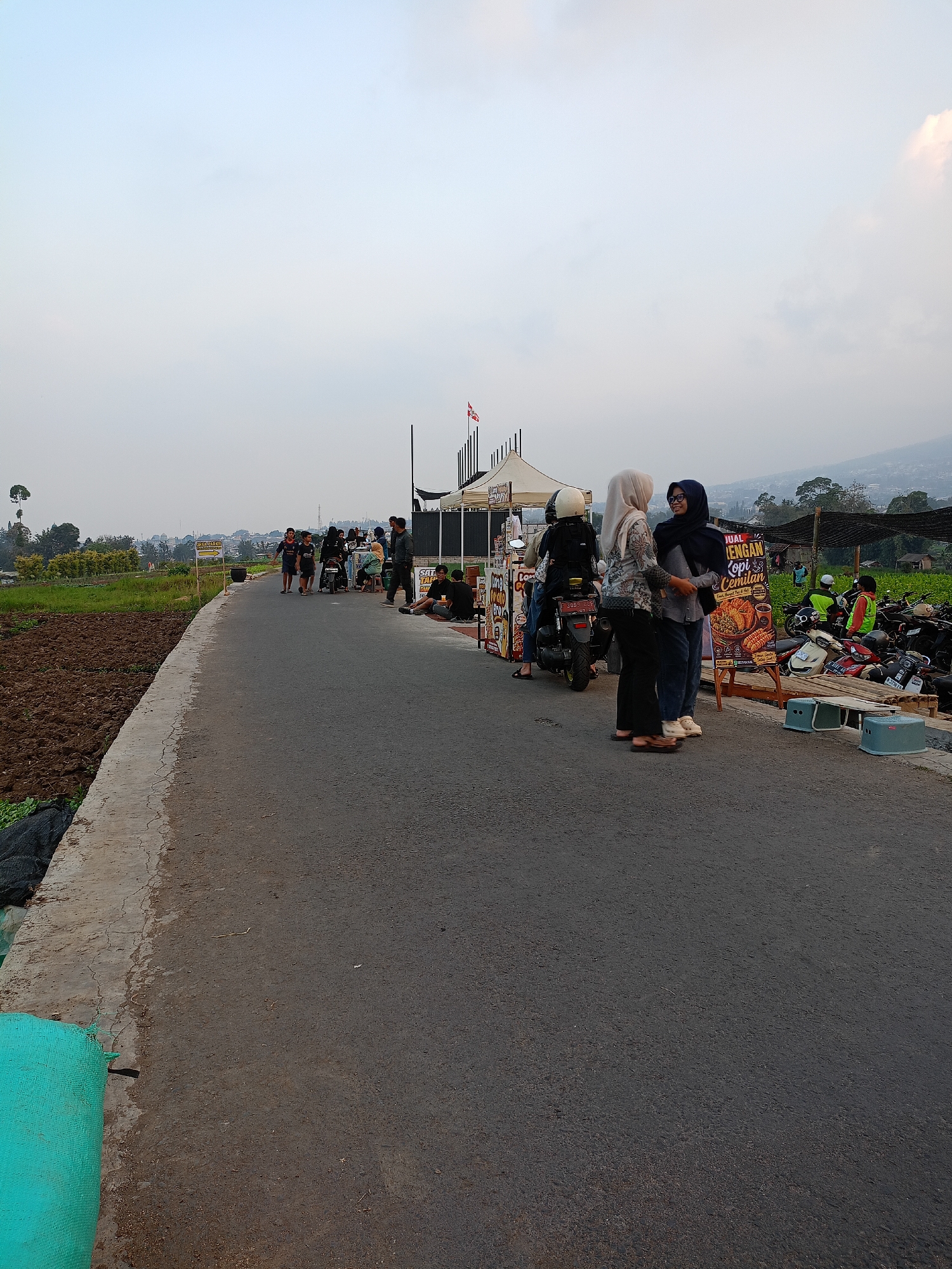 A group of people are gathered along a paved road in a rural or semi-rural area of Batu, Indonesia. The road appears to be a main thoroughfare, with a cultivated field to the left and a parking area or market setup on the right. Several motorcycles are parked on the right side of the road, suggesting a place of business or a gathering spot.
On the right side of the road, there are temporary market stalls or food vendors with tents and signage. One sign, in Indonesian, advertises "KOPI & CEMILAN" (Coffee & Snacks), with a picture of food. Two women in hijabs are standing in front of this stall, engaged in conversation. Another person, wearing a helmet and a black jacket, is sitting on a motorcycle nearby. Further down the road, more people are visible, some walking and some gathered around the stalls.
To the left of the road, a large area of tilled soil suggests agricultural activity. In the background, there are trees, buildings, and hills, characteristic of the landscape in Batu, which is known for its cooler climate and agricultural produce. The sky is overcast, with a pale, diffused light. A flag, possibly the Indonesian flag, is visible on a pole above one of the tents. The scene depicts a casual gathering and commercial activity in an outdoor setting.
