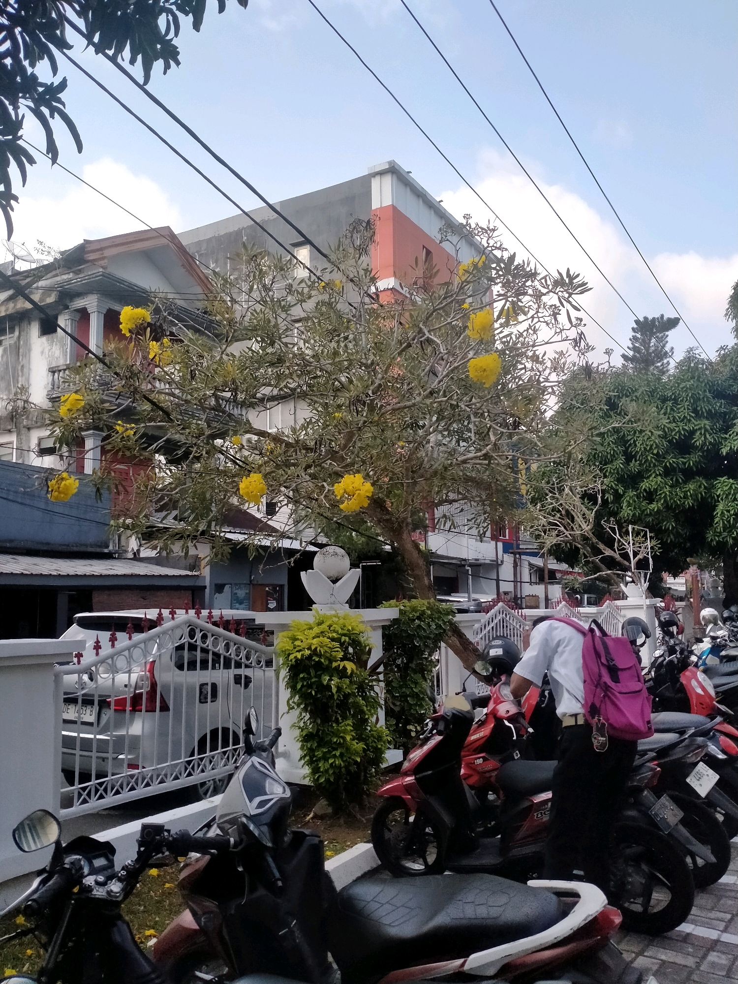 A person wearing a white shirt, black pants, and a pink backpack is standing next to a row of parked motorcycles, adjusting something on the handlebars of the closest motorcycle. The person is facing away from the camera. The motorcycles are parked on a paved surface next to a white fence. Beyond the fence, a white car is parked in front of a building. In the background, there is a tree with yellow flowers and several multi-story buildings. Overhead, power lines are strung across the sky. The scene appears to be in a residential or commercial area of Ambon, Indonesia, possibly on a street or in a parking lot.