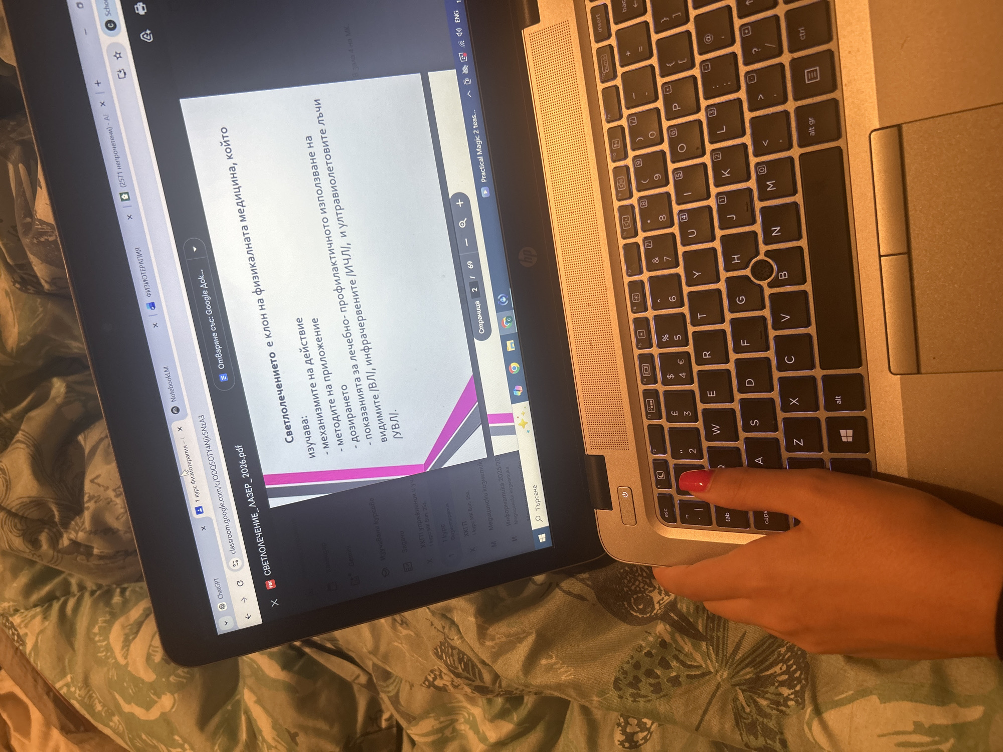 The image shows a person using a laptop. The screen displays a document related to physiotherapy, specifically phototherapy and laser treatment. The text on the screen is in Bulgarian. The laptop has a silver-colored keyboard and is resting on a surface with a patterned cover. A hand with pink nail polish is positioned near the keyboard's lower-left corner, apparently about to press a key or use the touchpad. The laptop is displaying a web browser with several tabs open, including Google Classroom, ChatGPT, and a PDF document related to phototherapy.