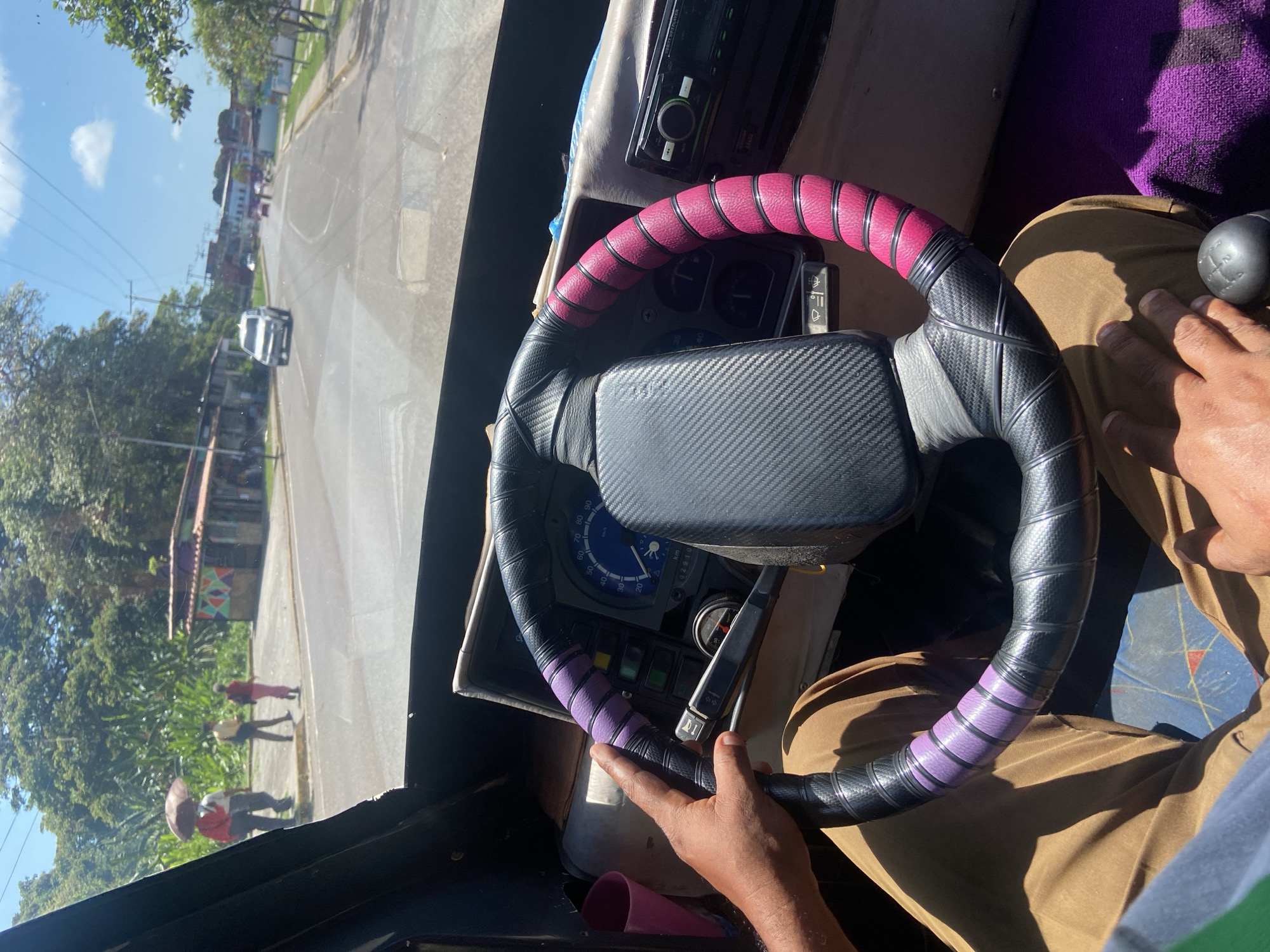 **Scene:** View from inside a vehicle, likely a bus, showing the steering wheel, dashboard, and a partial view of the road and cityscape outside.
**Location:** San José de Barlovento, Venezuela. The buildings and environment visible through the windshield suggest a densely populated area within the town.
**Objects:** The steering wheel is wrapped with a black carbon fiber textured material, accented with pink and purple bands. Dashboard with a speedometer. Car radio. A gear stick. The exterior shows a street with buildings, trees, power lines, and a white car.
**People:** The hands of a person are on the steering wheel. Pedestrians can be seen in the distance on the sidewalk.
**Action/Interaction:** The person is driving the vehicle, presumably through the streets of San José de Barlovento.