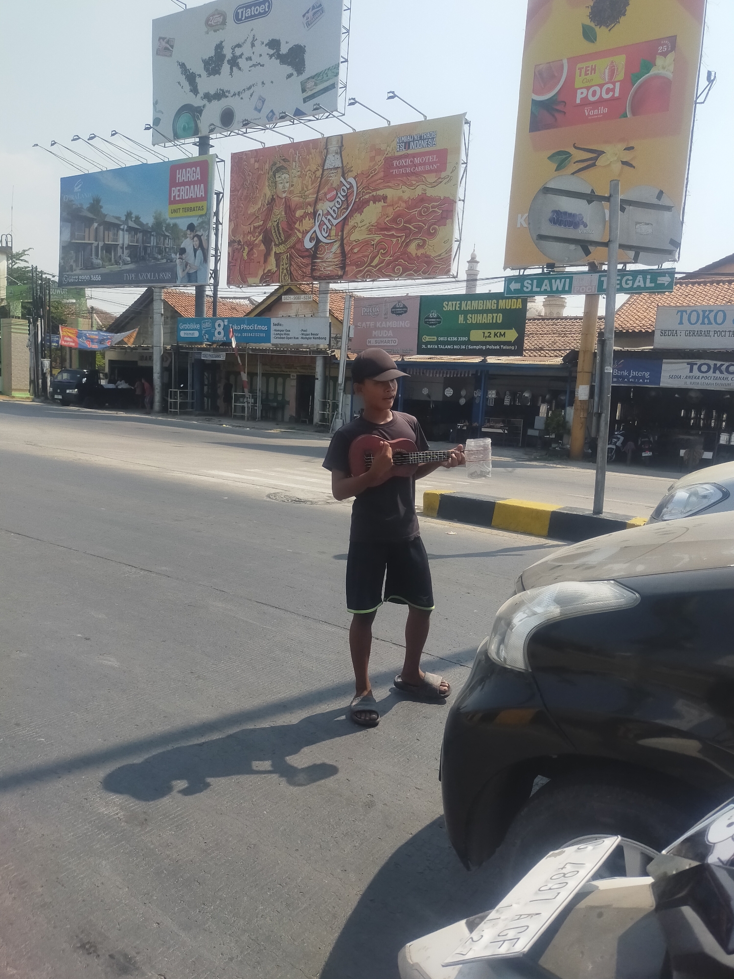 In Lemahduwur, Indonesia, a young boy stands on the side of a road, holding a ukulele and looking towards the camera. He is wearing a dark t-shirt and shorts, and sandals. Next to him is a plastic bottle, likely for collecting money. In the foreground, the hood of a black car is visible, partially obscuring the boy's legs. Behind him, the street is lined with buildings and businesses. Several large billboards are prominently displayed overhead, advertising various products and services, including real estate, tea, and food. The sky is clear and bright. The overall scene depicts a street view with a street performer and commercial advertisements.