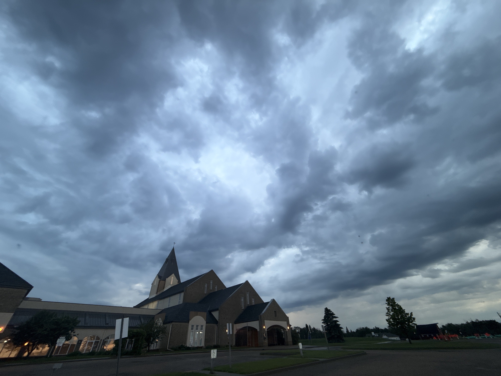 An exterior, low-angle, wide shot depicts the dark, overcast sky above a church building in Stony Plain, Canada. The sky is filled with dense, grey clouds that partially obscure the light. The church building, constructed of brick, features a tall, pointed steeple topped with a cross. A portion of a larger, modern wing with large windows is visible to the left of the main church structure. The foreground consists of a paved area and a grassy verge. There are no visible people in the image. The overall atmosphere is somber due to the weather conditions.