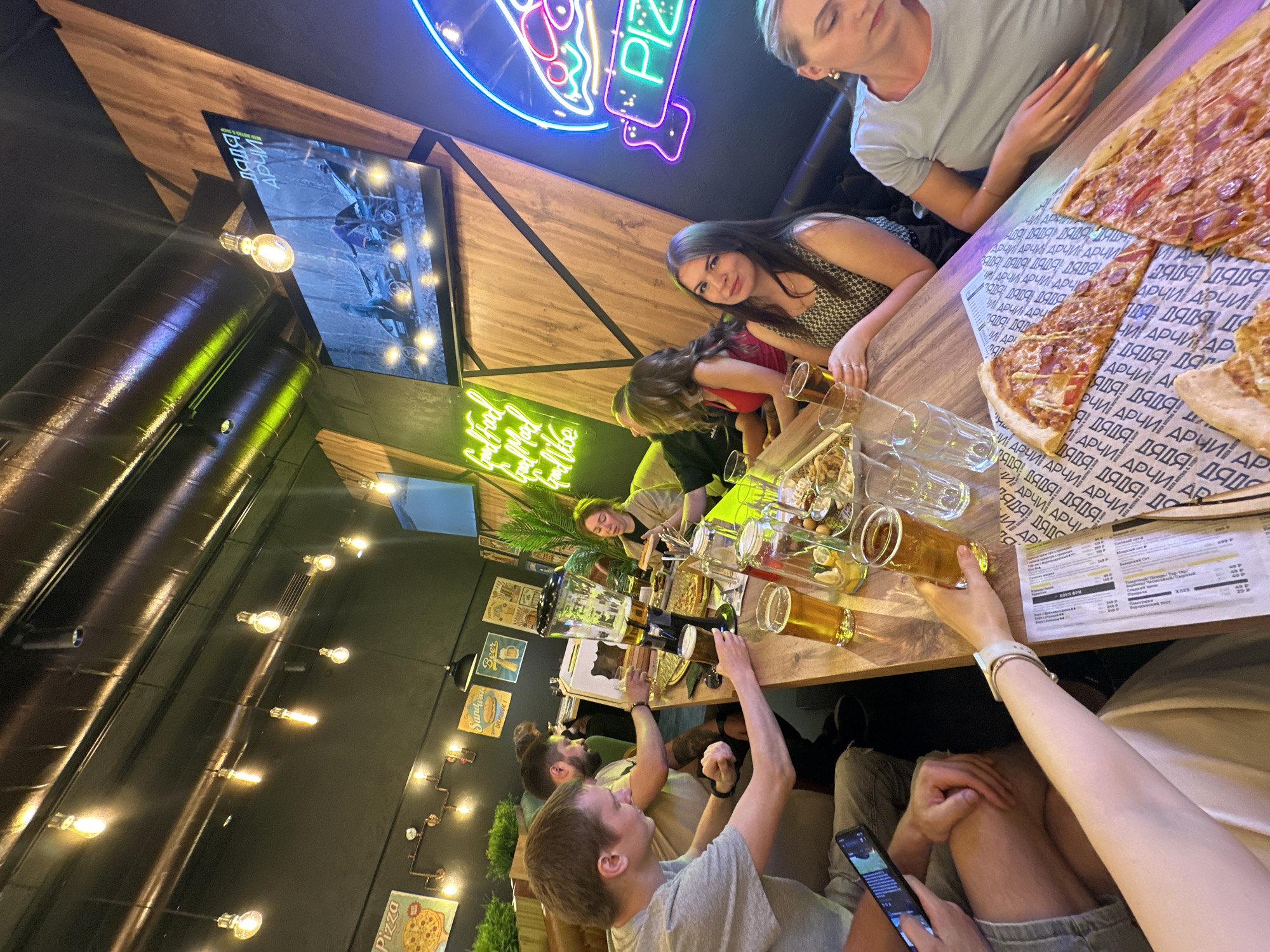 Here's a detailed description of the image:
**Scene:** The image captures a group of approximately 10 people gathered around a long wooden table in what appears to be a pizzeria in Perm, Russia. The setting has a modern, slightly industrial vibe, evidenced by exposed ceiling elements and decorative lighting.
**People:** The group consists of both men and women, varying in age. Some are engaged in conversation, others are looking at their phones, and some are interacting with the food and beverages on the table. There's a general sense of conviviality and shared enjoyment.
**Subjects:** The primary subject of the image is the social gathering itself, with secondary subjects including the food and drinks on the table and the restaurant's decor.
**Objects:** Key objects include:
* A large pizza served on paper with Russian text "ДЯДЯ АРЧИ" (Daddy Archie) which might refer to the restaurant's name.
* Several beer glasses filled with beer.
* Beer draft tap with a beer tower.
* A smartphone being held by one of the individuals.
* Decorative lighting fixtures.
* A television screen displaying a movie or show.
* Neon signs on the wall, including one shaped like a pizza slice and one that says "Good Food, Good Time, Good Vibes."
* Menus on the table.
**Actions/Interactions:** People are seen:
* Raising hands to pour beer.
* Drinking beer.
* Talking and laughing.
* Using their phones.
**Environment:** The environment is characterized by:
* A modern restaurant setting with wooden wall panels.
* Industrial-style lighting fixtures hanging from the ceiling.
* Decorative plants and artwork.
* A neon pizza sign adding a playful touch.
* A television screen displaying content.