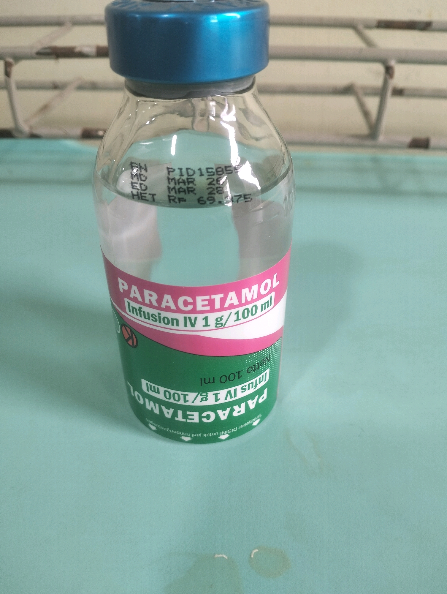 A clear glass bottle with a blue cap contains a pale liquid and is labeled with "PARACETAMOL Infusion IV 1 g / 100 ml". The bottle is standing on a light blue surface. In the background, a metal rack is visible. The image is a close-up of the bottle, with the label partially obscured by the bottle's curvature. The scene appears to be in a medical setting. There is no indication of the location in the city of Air Jamban, Indonesia.