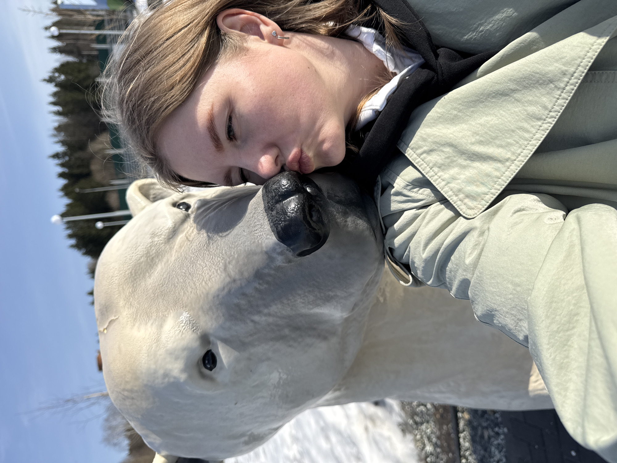 A young woman with blonde hair and a silver heart earring is kissing the black nose of a white, non-living polar bear statue. The woman is wearing a light green jacket over a black hooded sweatshirt and a white shirt. Patches of snow are visible at the bottom of the frame. Trees and a clear blue sky are in the background. The location is Perm, Russia.