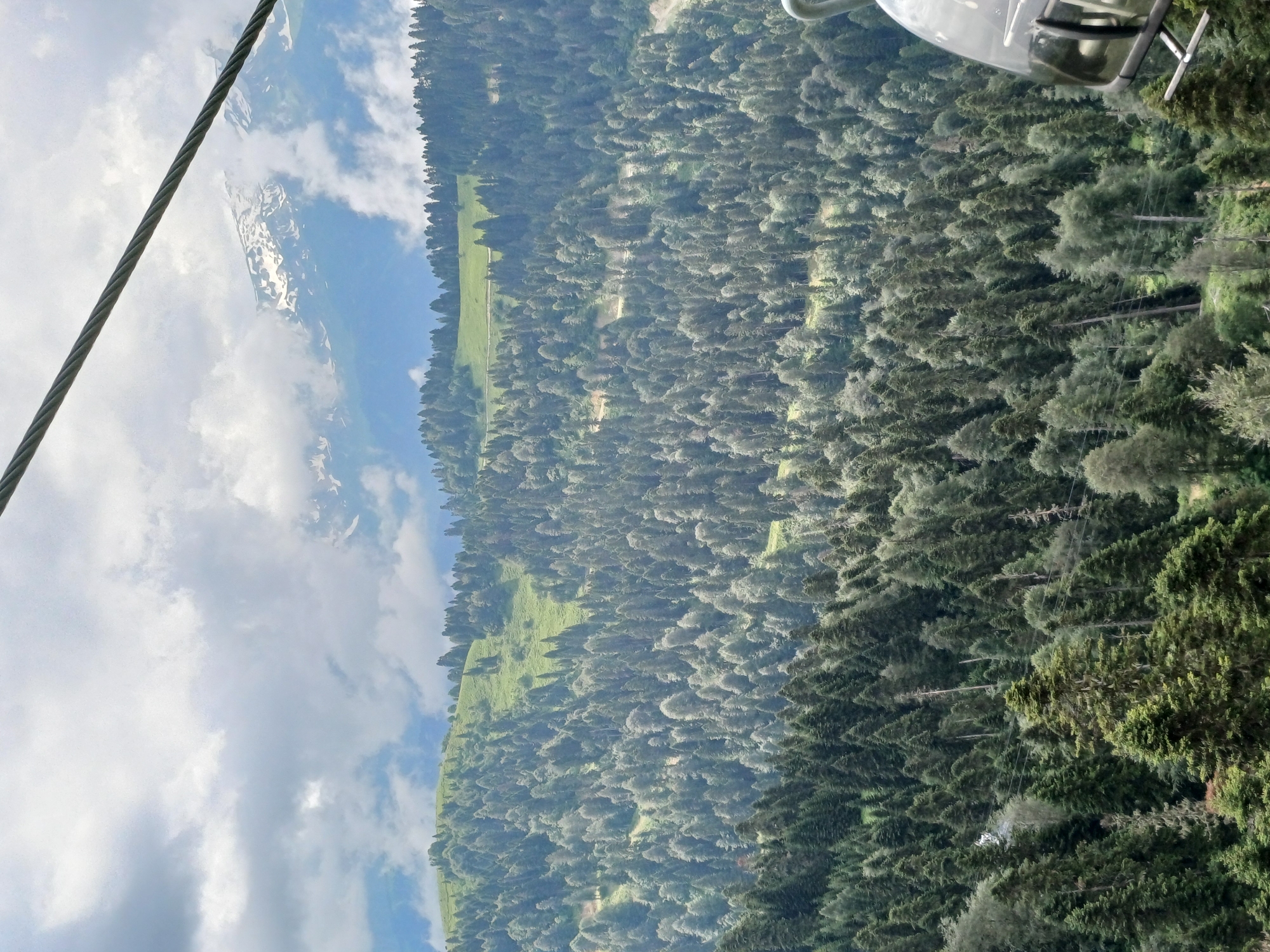 A cable car moves through a mountainous landscape covered in dense evergreen forests. The sky is partly cloudy with patches of blue visible. In the distance, snow-capped mountains are partially obscured by clouds. The cable car's cabin is partially visible on the right side of the image. The scene appears to be from an elevated perspective, likely taken from within another cable car or a similar vantage point. The location is Estosadok, Russia, a ski resort town in the Caucasus Mountains.