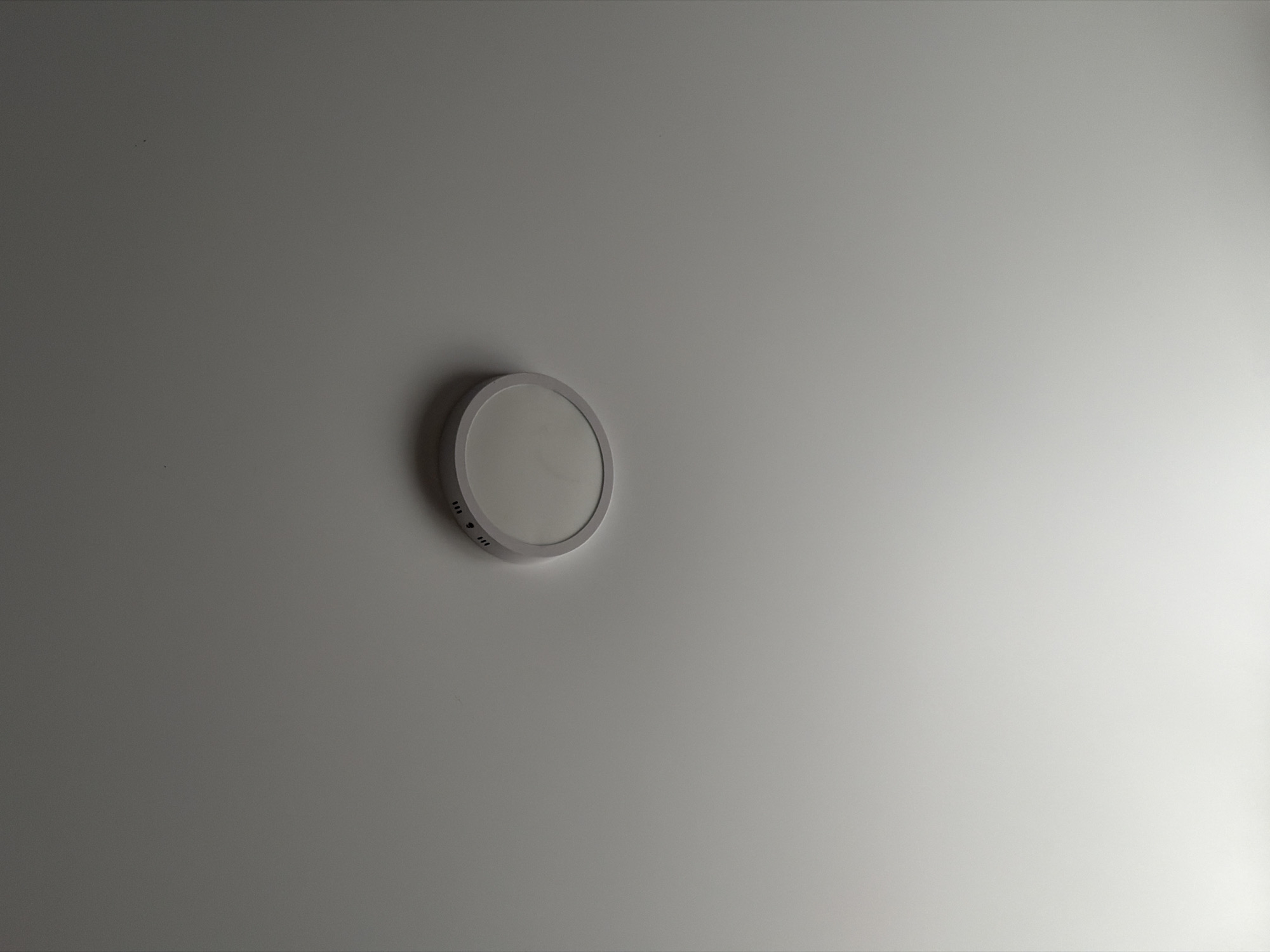 The image shows a modern, round LED ceiling light fixture mounted on a plain, off-white ceiling. The fixture has a thin, metallic rim and a frosted diffuser. There are small, dark specks visible on the ceiling, which could be dust. The lighting appears to be from the fixture itself, casting a soft glow on the surrounding ceiling. The perspective is from below, looking upwards at the fixture. The location in the city of Strășeni, Moldova, is not discernible from the image alone.