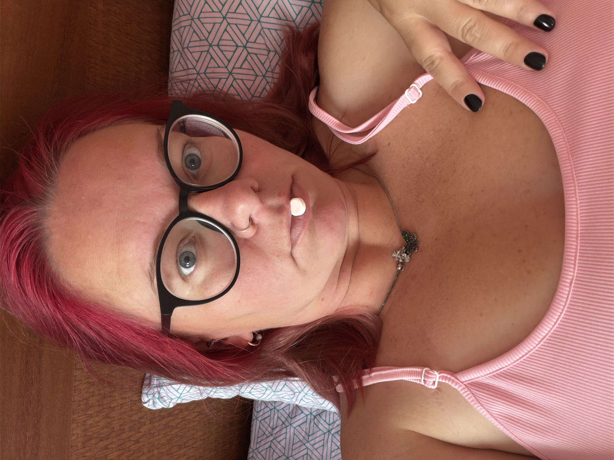 A woman with bright pink hair and black-rimmed glasses is lying on a bed, looking up at the camera. She has a small white pill or object balanced on her lips. She is wearing a pink tank top and a necklace with a butterfly pendant. The background shows a wooden headboard and a pillow with a geometric pattern. The scene is indoor and appears to be a bedroom. There is no indication of the location being in Chaikovsky, Russia.