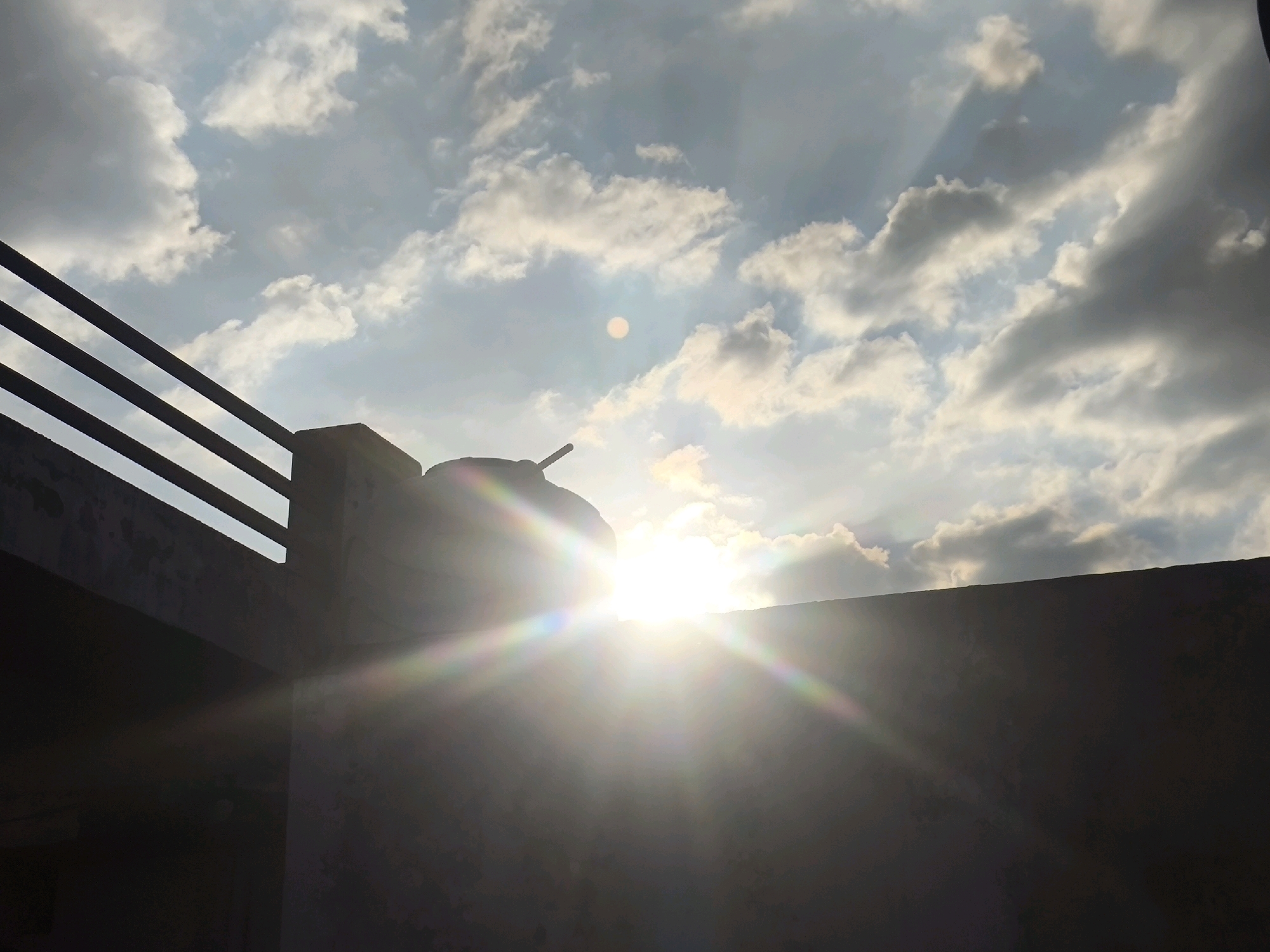 The image depicts a view of the sky from a rooftop in Chhajpur Kalan, India. The sun is intensely bright, creating a lens flare that obscures much of the detail. Wispy white clouds are scattered across a pale blue sky. In the foreground, silhouetted against the bright sky, is a rooftop structure. This structure includes a railing with horizontal bars and a round water tank. The lower portion of the image is a dark, unlit area, likely the side of a building. The overall scene captures a sunny day with a dramatic sunburst effect.