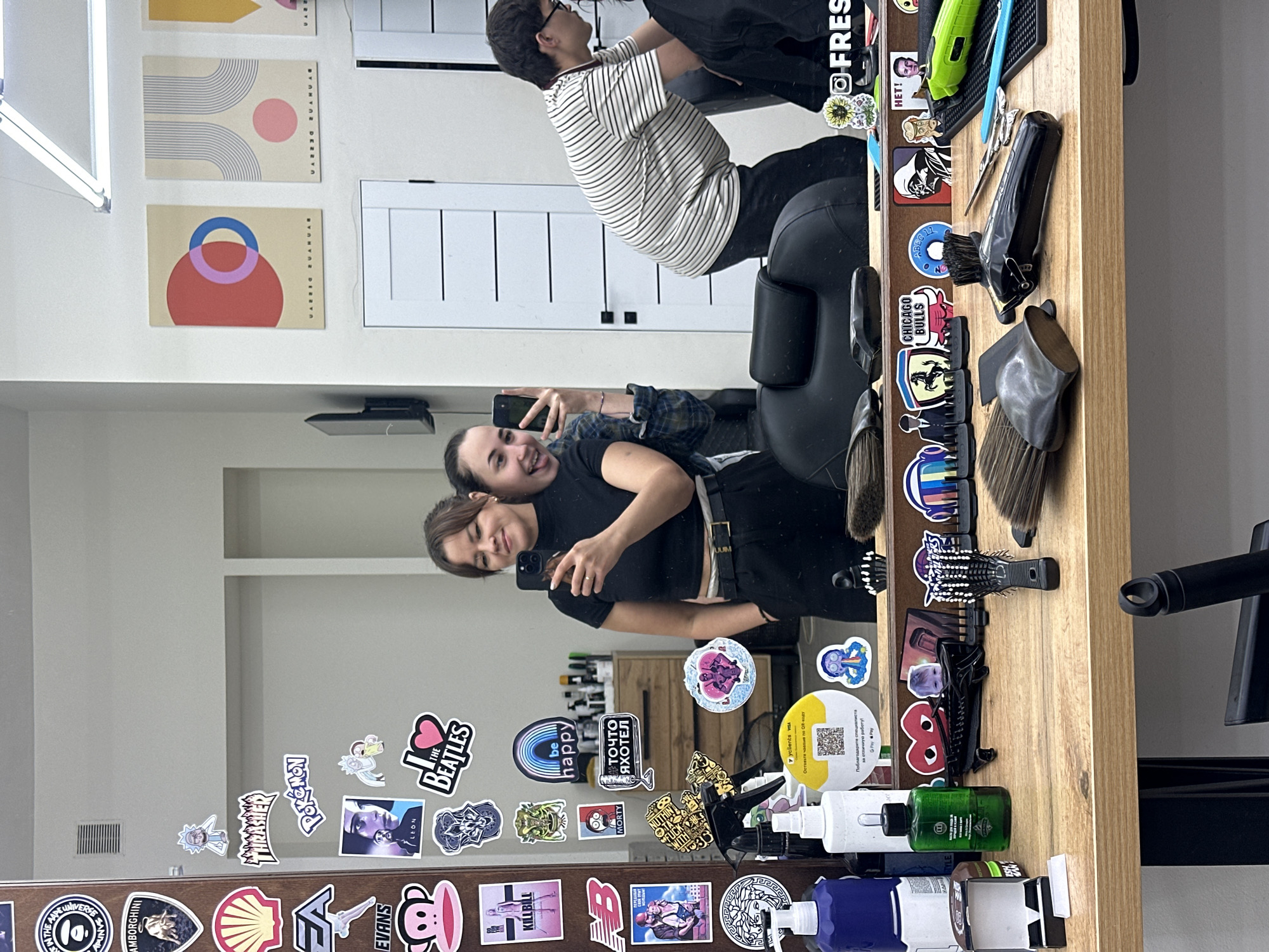 The image depicts a mirror reflection in what appears to be a barbershop. Two young women are taking a selfie in the mirror. The woman in the foreground on the left is wearing a black crop top and black pants, with her arm raised to take the photo. The woman behind her, on the right, is wearing a plaid shirt and sticking out her tongue. Both women have their phones out.
The background shows a wooden counter with various barbering tools and products, including brushes, scissors, clippers, and bottles of hair products. Numerous stickers are adhered to the mirror and the wall behind the counter, featuring brands, characters, and pop culture references. On the wall, there are also framed minimalist art prints. A person with glasses, wearing a striped shirt, is visible in the reflection behind the counter, appearing to be working with a client. The environment suggests a modern and trendy barbershop. The context of Chaikovsky, Russia, is noted, implying this scene is within that city.