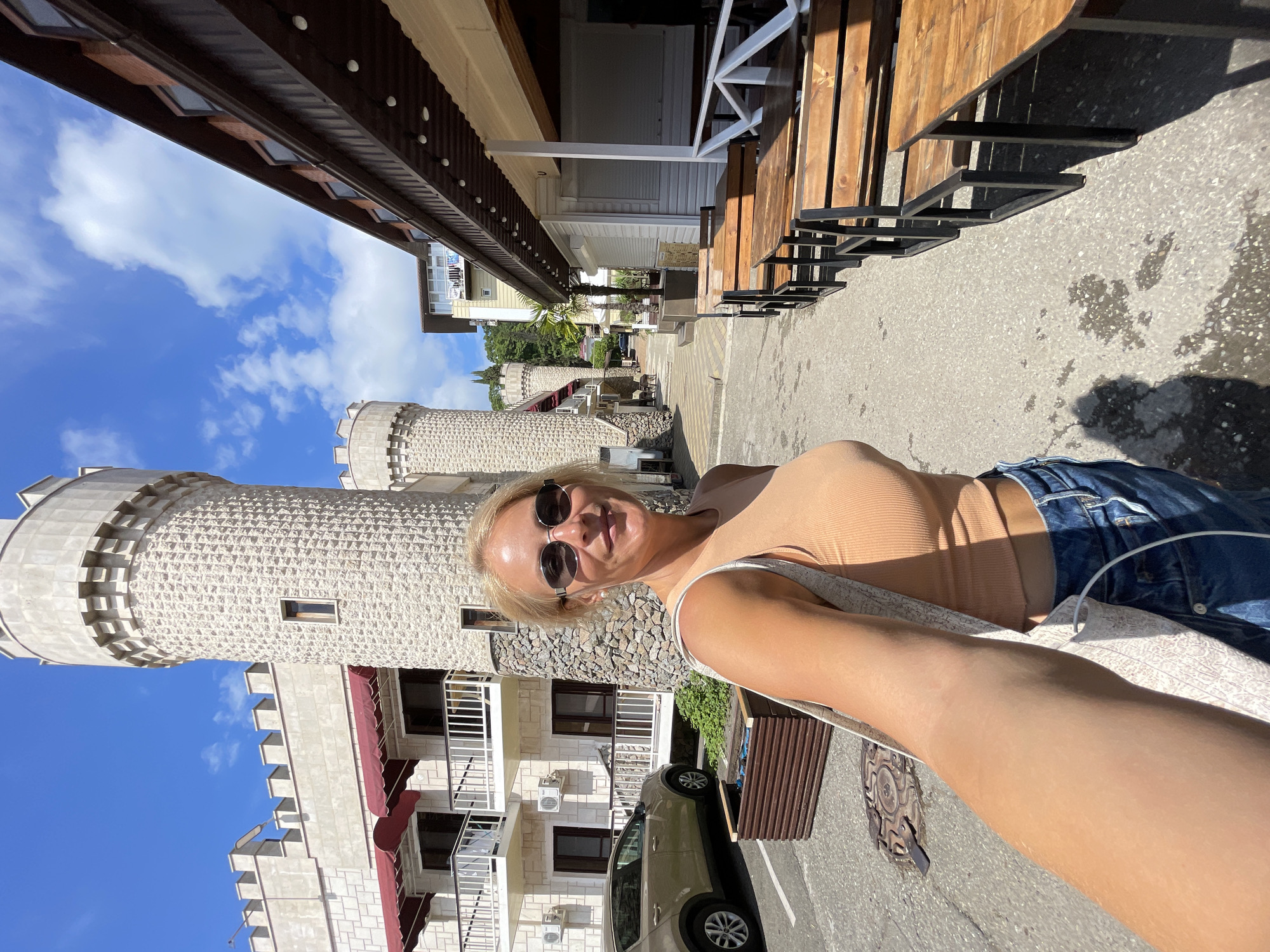 A woman with blonde hair and sunglasses is taking a selfie in front of a building that resembles a castle, located in Sochi, Russia. She is wearing a light orange tank top and blue jeans. In the background, there are two tower-like structures with stone cladding and crenellations. To the right of the woman, there is an outdoor seating area with wooden tables and benches, likely belonging to a cafe or restaurant. The street is paved, and a car is parked to the left of the woman. The sky is blue with scattered clouds. The architectural style suggests a resort or tourist area, possibly along the coast.