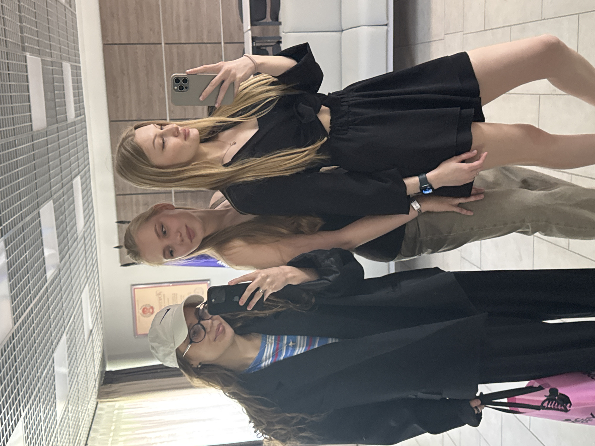 Three women are taking a selfie in what appears to be a mirrored area, possibly a shopping center or public space. The woman on the top is holding a phone and wearing a black dress. The woman in the middle has blonde hair and is wearing a shirt and light brown pants. The woman on the bottom is wearing a white baseball cap, glasses, a striped shirt, and a black jacket. A pink shopping bag is visible in the bottom right corner. The background includes a wall with square tiles, a piece of furniture that appears to be a white sofa, and a window with what looks like window blinds. The location is Perm, Russia, identified by a certificate with the city's emblem on the wall behind the women.