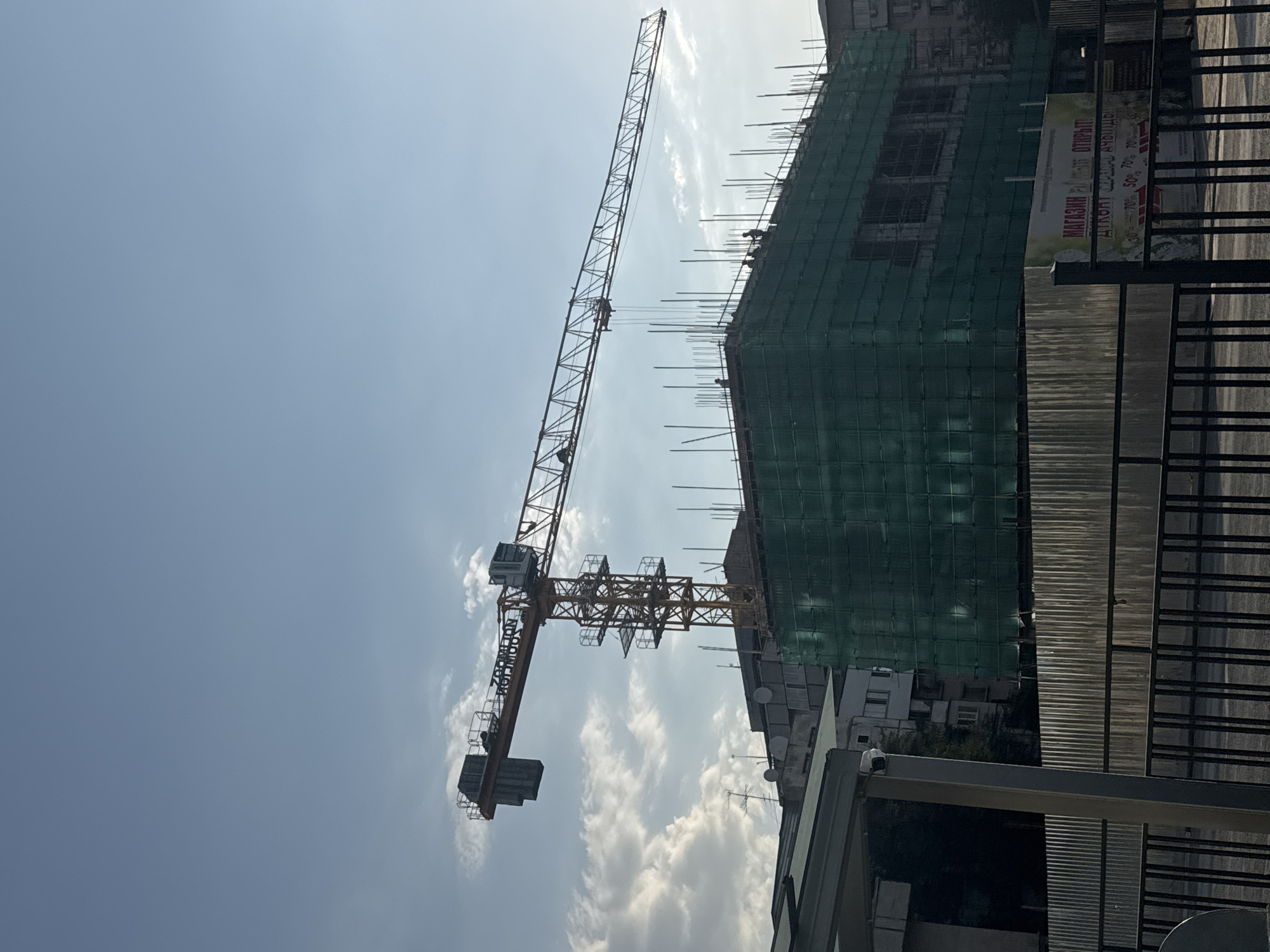 A yellow construction crane with the lettering "ZOOMLION RUJIINDUO" is positioned over a building under construction. The building is partially covered in green netting, and rebar protrudes from its upper levels. Several individuals, appearing as small silhouettes, are visible on the top of the building. In the foreground, a corrugated metal fence and a black metal fence are present. A sign on a white banner in the lower right corner reads "МАГАЗИН РАБОТАЕТ! ОТКРЫТ!," indicating that a store is open. Text below this appears to be advertising discounts, showing "30%, 50%, 70%". The scene is set against a clear blue sky with some scattered clouds. This image was taken in Bishkek, Kyrgyzstan.