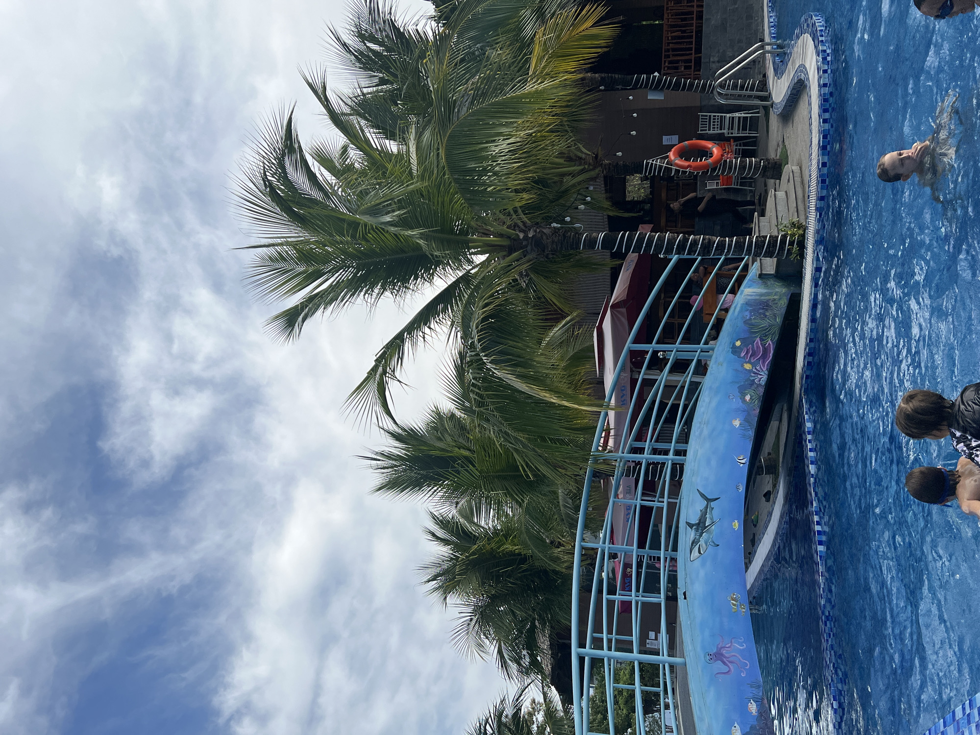 The scene, located in Nam Nha Trang Ward, Vietnam, features a swimming pool adjacent to a structure with a red life preserver hanging on the exterior. Palm trees are visible, with some branches extending near the building. A blue fence painted with sea creatures borders part of the pool. Two children are in the water, and an adult is swimming in the deeper end. The sky is partially cloudy.