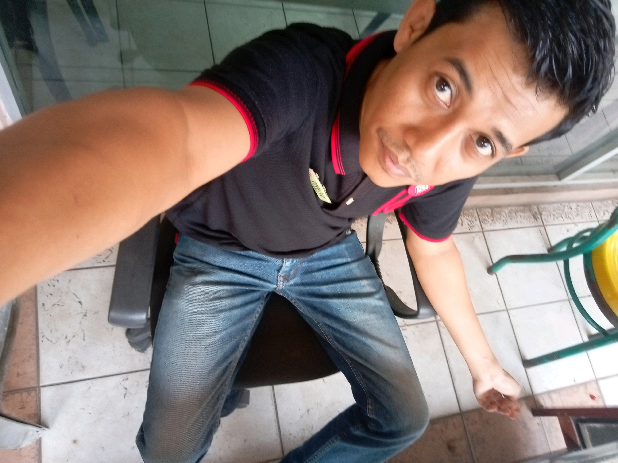 The image is a selfie taken by a person sitting in a chair. The person is wearing a black polo shirt with red trim and blue jeans. The person's left arm is extended towards the camera, and their face is looking at the camera. The background shows a tiled floor and a green chair with a yellow object on it. The location is identified as Batam, Indonesia.