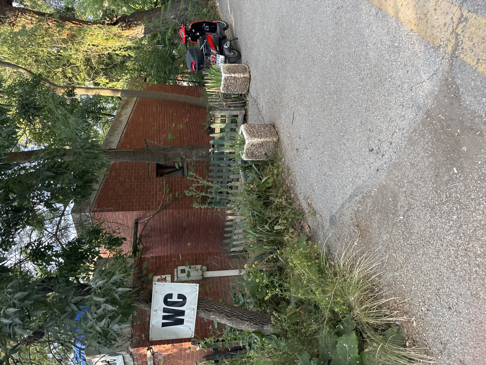A red and black scooter is parked on an asphalt road next to a brick building with a sign that says "WC". Two square planters with greenery sit on the ground near the scooter. A wooden fence separates the planters from a grassy area with a tree. The scene is located in Gölbaşı, Turkey.