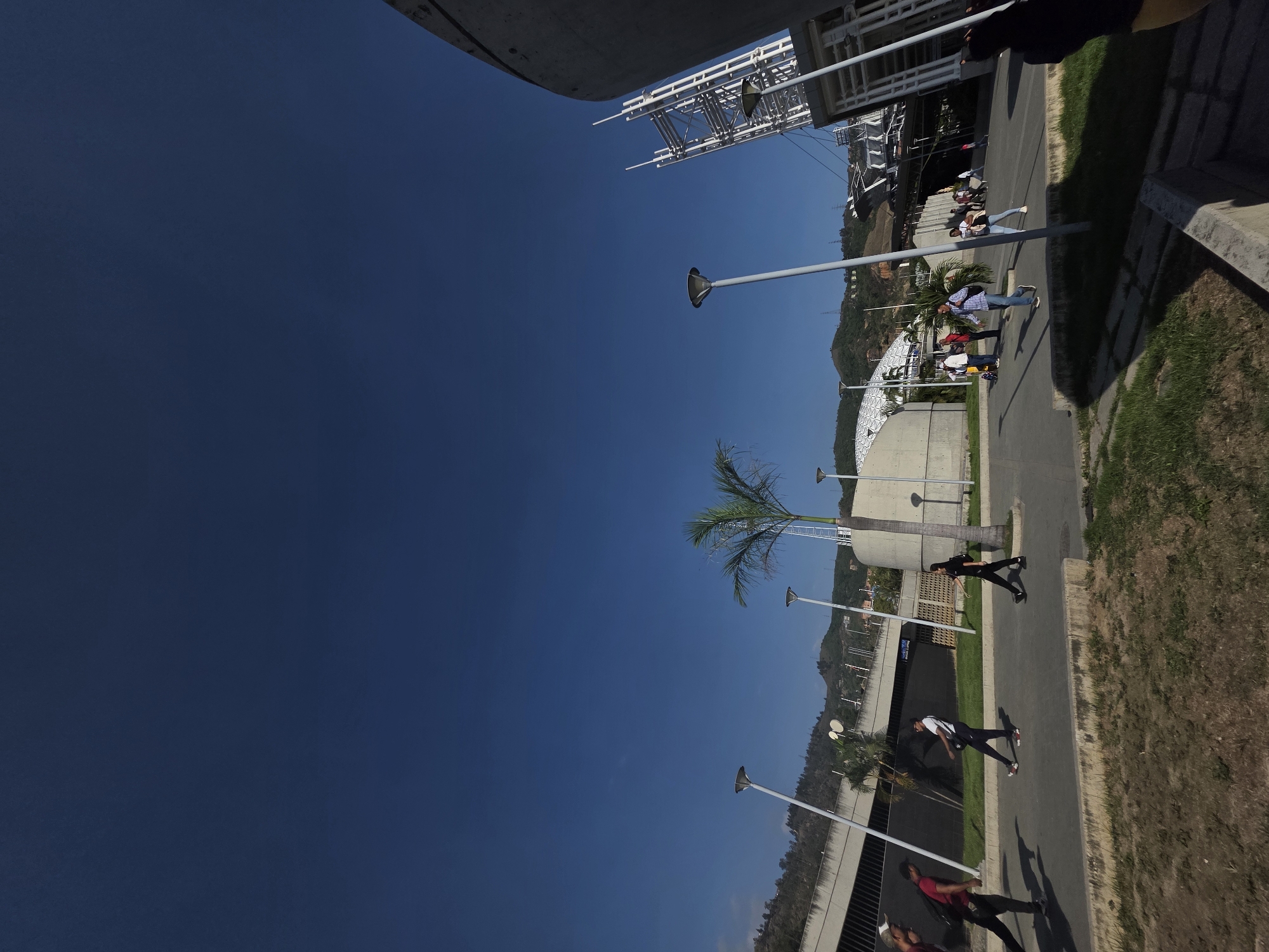 The image depicts an outdoor scene in Caracas, Venezuela. A clear blue sky dominates the left portion of the frame. A sidewalk runs along the right side of the image, with pedestrians walking on it. Street lamps line the sidewalk. A palm tree is visible amidst the street lamps. Buildings and vegetation are on the hillside in the background.