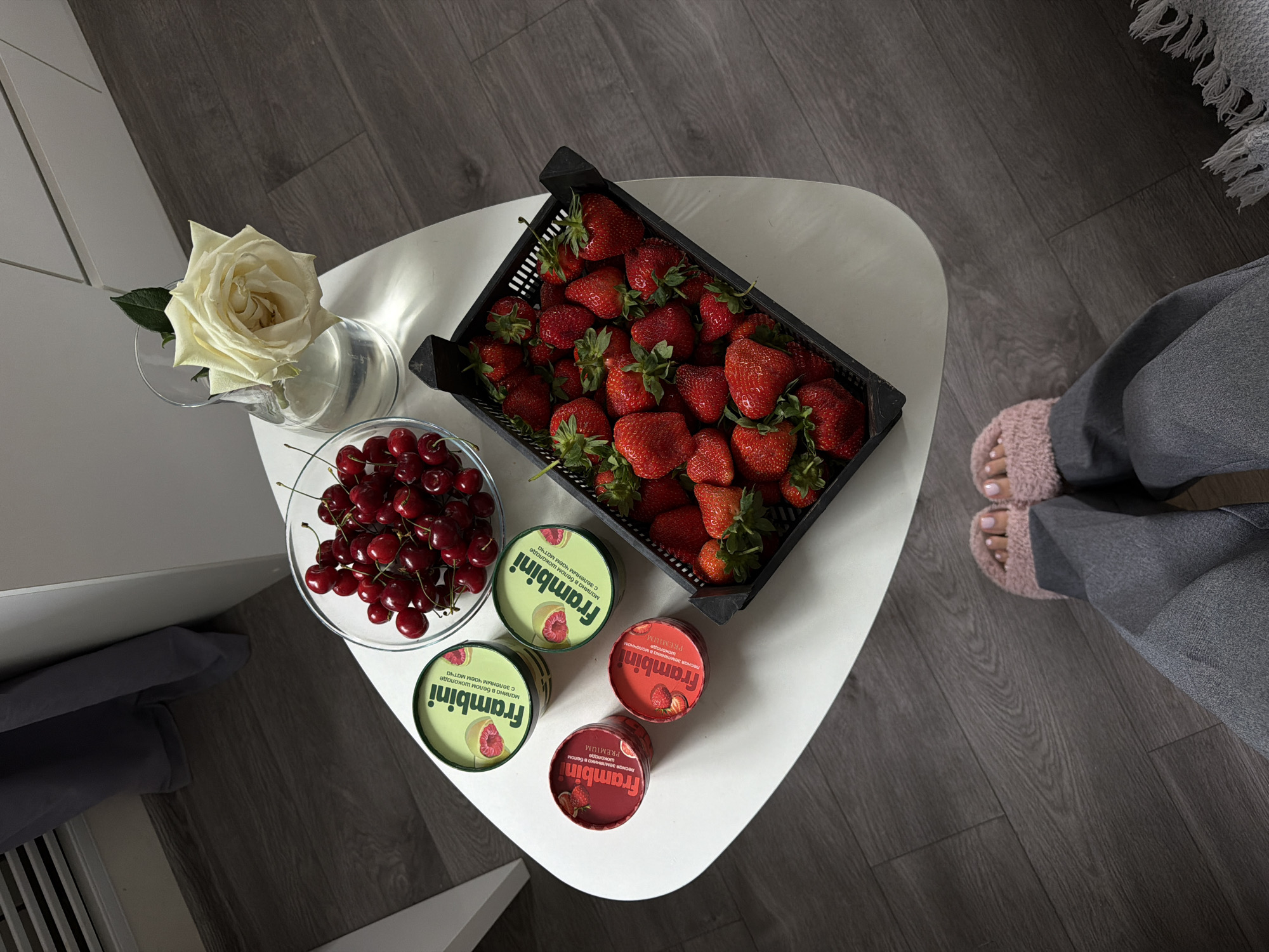 A high-angle shot captures a white, triangular coffee table positioned on a gray wood-patterned floor. On the table, a black plastic crate overflows with fresh strawberries. Adjacent to the strawberries, a glass bowl contains a pile of cherries. Next to the bowl, four containers of "Frambini" ice cream are arranged. To the left, a white rose is placed in a glass pitcher filled with water. To the right of the table, a person wearing gray pants and pink furry slippers is visible. The setting suggests an indoor environment, possibly in Kazan, Russia, given the brand of ice cream.