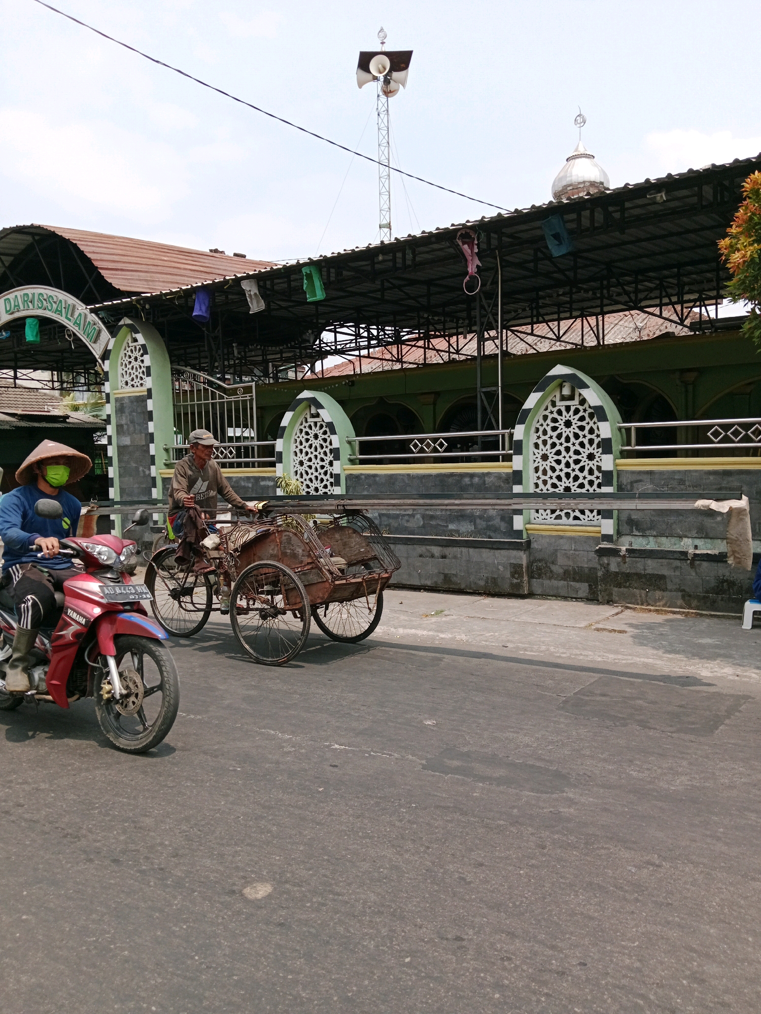 In Cemani, Indonesia, on a paved road in front of a building with a domed roof and a large awning, two people are visible. On the left, a person wearing a blue long-sleeved shirt, black pants, boots, a wide-brimmed conical hat, and a light green face mask rides a red and silver Yamaha motorcycle. The motorcycle's license plate is partially visible. To the right of the motorcycle, an older man wearing a light brown long-sleeved shirt, dark pants, and a cap is pedaling a traditional pedicab. The pedicab, which is made of weathered metal and wood, has two large spoked rear wheels and a single spoked front wheel. It appears to be empty. The building behind them has green walls and black and white decorative elements around arched windows. A tall metal tower with speakers is visible above the awning on the left side of the frame. Several colorful cloth banners or decorations are hanging from the awning. The sky is overcast.