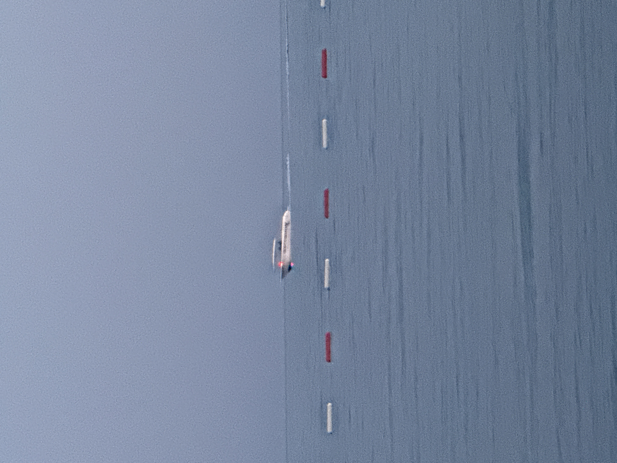A speedboat is cutting through the calm, grey waters of the Adriatic Sea off the coast of Boreti, Montenegro. The boat is white with red and blue accents and appears to be moving at a moderate speed, leaving a visible wake. In the foreground, a line of buoys, alternating between white and red, mark a boundary in the water. The sky is overcast and a muted blue-grey, creating a somewhat hazy and serene atmosphere. The image captures a typical coastal scene with the sea, sky, and markers delineating a swimming or boating area.