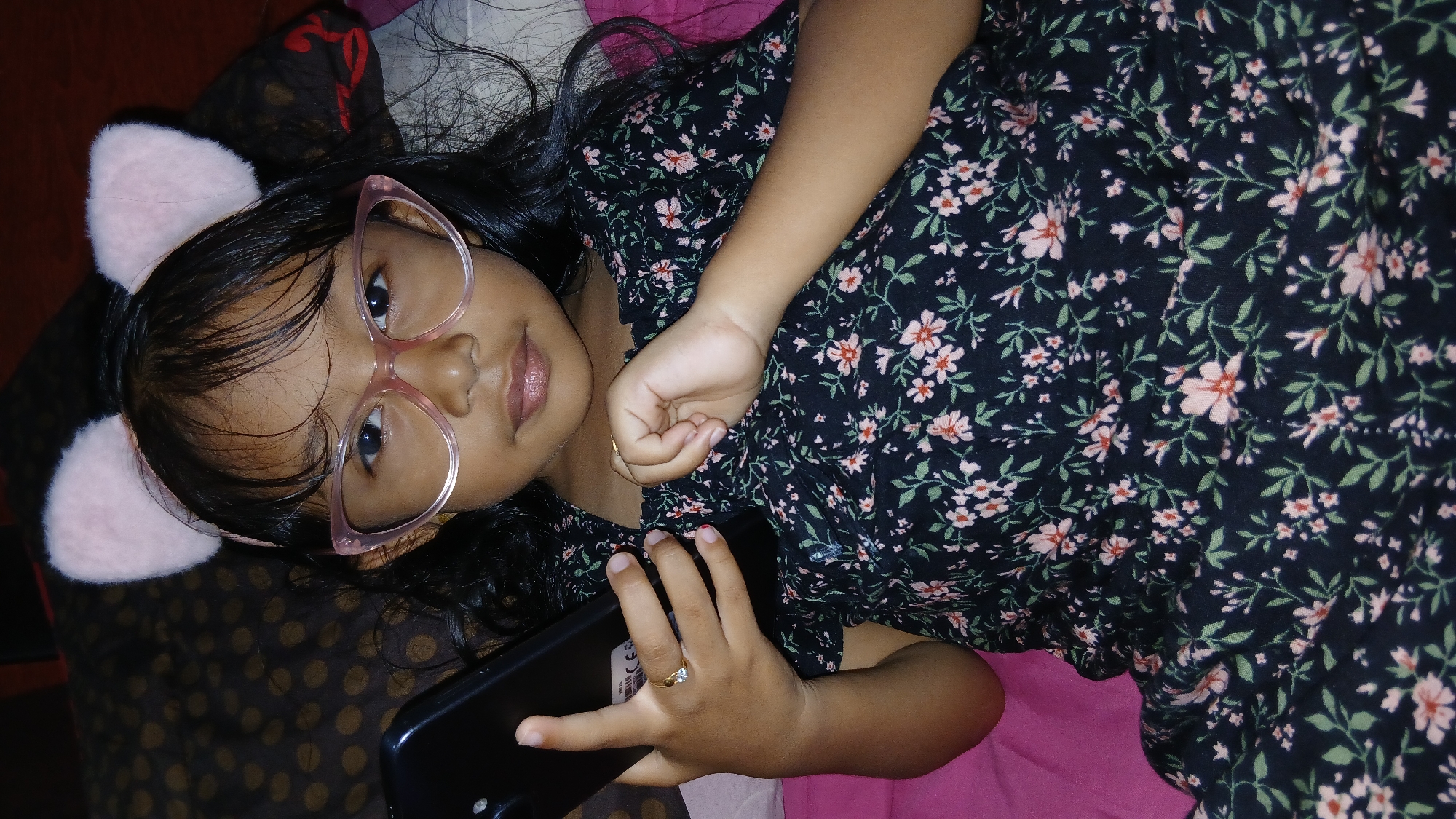 A young girl with long, dark hair and pink cat ears on a headband is lying down, holding a smartphone in her left hand. She is wearing pink, cat-eye glasses and a dark, floral-print dress. Her right hand is resting on her chest. The background is a bed with a patterned blanket and a pink sheet. The scene is indoors.