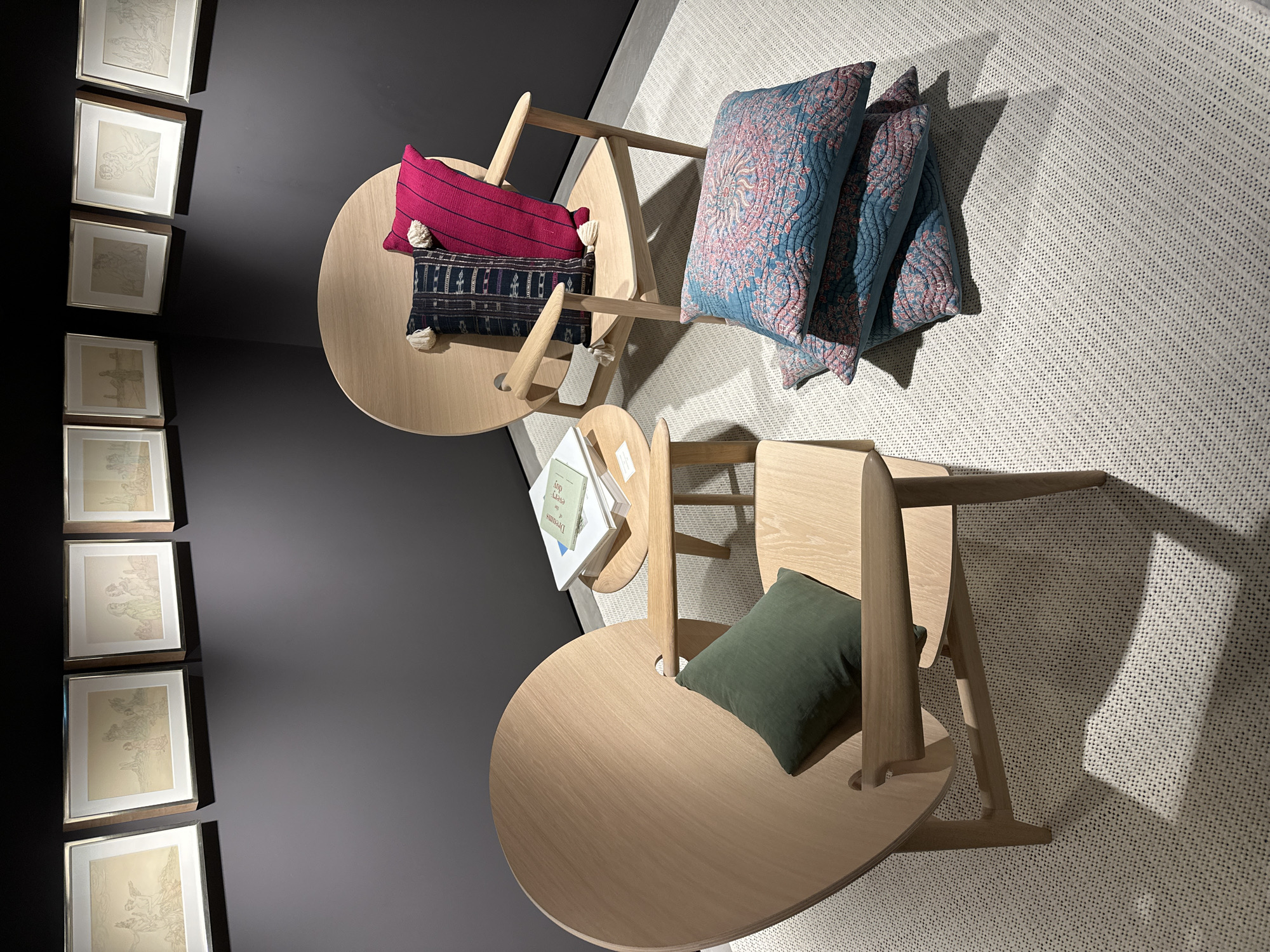 This media file captures an indoor scene, likely a contemporary furniture showroom or a curated living space, located in London, United Kingdom. The central subjects are two light-colored wooden chairs, each featuring a unique, modern design with a circular seat and an integrated small, round table.
No people are visible; the scene is static, showcasing the furniture and decor. On the chair positioned further away, two rectangular pillows are placed: one is bright magenta with black stripes and white tassels, and the other is dark indigo with a woven ethnic-style pattern and white tassels. On the chair closer to the camera, a single dark green rectangular pillow rests. Additionally, three turquoise-teal quilted pillows with pinkish-red floral or paisley patterns are stacked on the cream-colored, textured rug.
On the dark gray wall to the left, a vertical series of at least seven framed artworks, possibly etchings or sketches depicting human figures, are displayed. These pieces are set in light frames with white matting. On the small integrated table of the closer chair, a stack of books is visible, with titles such as "every day" and "dreams" partially discernible. The artificial lighting creates distinct shadows, highlighting the textures of the rug and the forms of the furniture. The overall aesthetic is minimalist, comfortable, and modern.