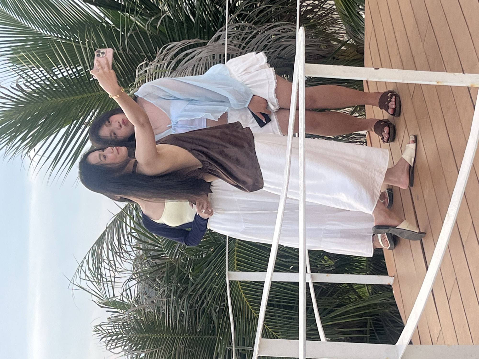 Two women are taking a selfie in Mũi Né, Vietnam. One woman, on the left, is wearing a light yellow camisole and a dark blue cardigan, while the other woman, on the right, is wearing a blue sheer blouse over a light pink top and a white skirt. The woman on the right holds a pink smartphone to take the photo. They are standing on a wooden deck, possibly a balcony or patio, with a white railing. Lush green palm fronds are visible in the background, suggesting a tropical or subtropical environment. The lighting appears to be natural, indicating daytime. The women's expressions are relaxed and happy, with one of them smiling. The overall mood is casual and cheerful, likely a vacation or leisurely outing.