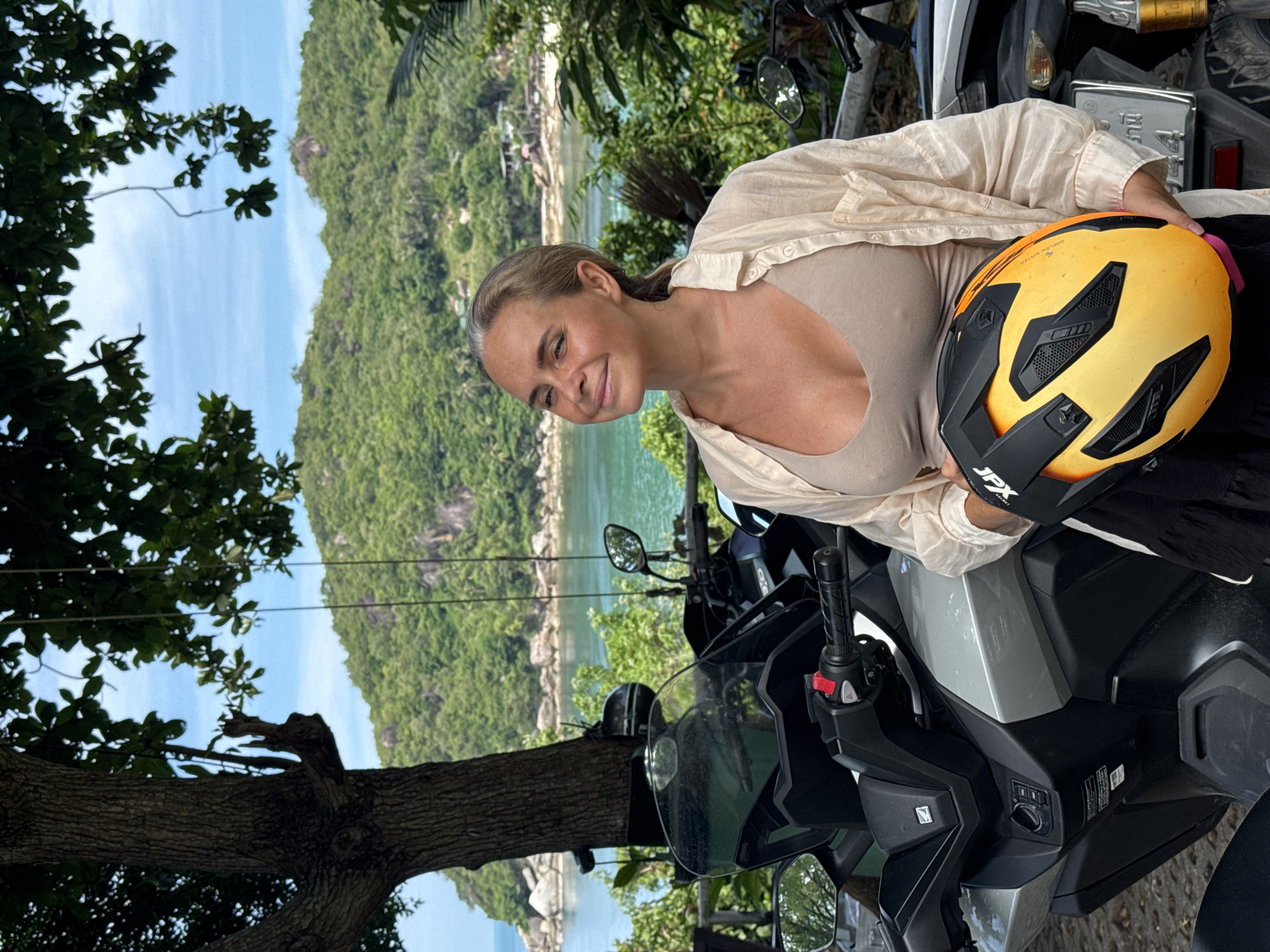 In a tropical coastal setting on Ko Samui District, Thailand, a woman stands next to a motorcycle, holding a yellow and black helmet. She is wearing a light beige tank top and an unbuttoned, light-colored long-sleeved shirt. Her hair is pulled back in a ponytail. The background features lush green hills, turquoise water, and a blue sky, with a large tree to the left of the frame. The motorcycle is dark-colored with a silver front panel and a prominent windshield. The license plate on the motorcycle is visible with Thai characters. The overall scene suggests a travel or leisure context.