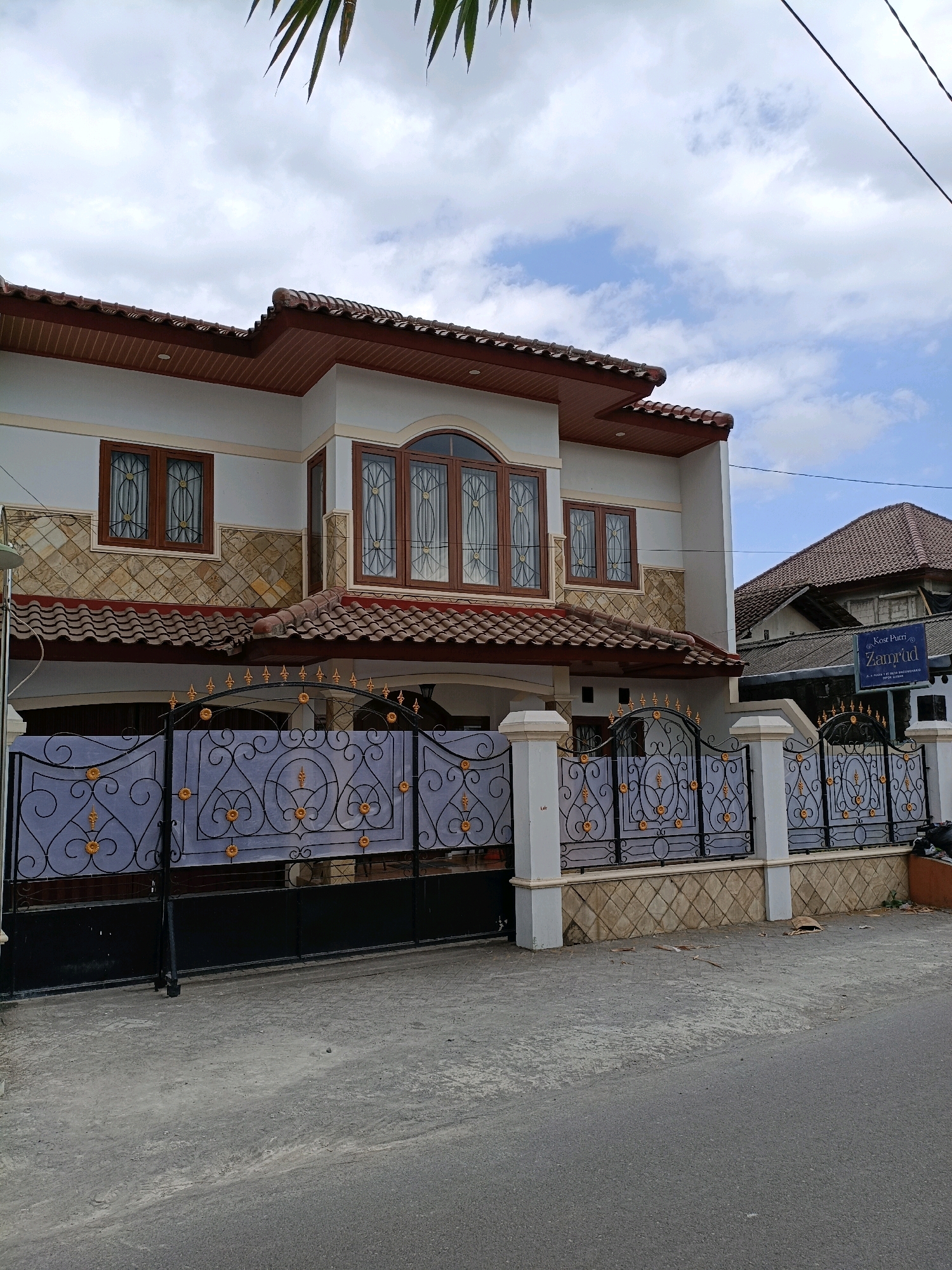 This image shows a two-story house with a white facade and a brown tiled roof. The house has multiple windows with brown wooden frames and decorative grills. A black metal gate with intricate golden scrollwork guards the entrance to the property, which is fronted by a paved driveway and sidewalk. To the right of the house, a blue sign with white lettering reads "Kost Putri Zamrud," indicating a boarding house for women. The sky above is partly cloudy, with hints of blue sky peeking through. The scene is likely located in Maguwoharjo, Indonesia, given the context.