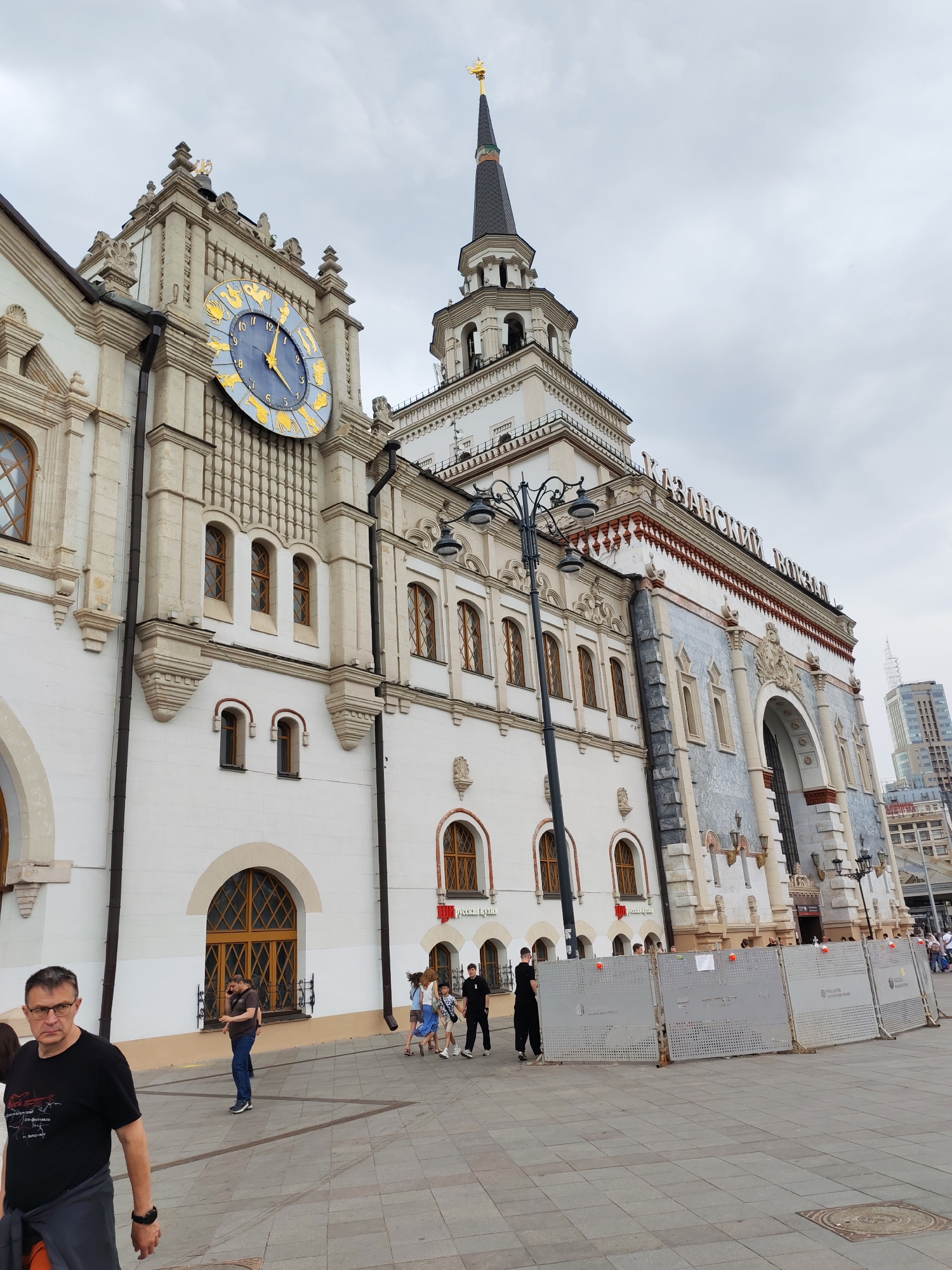 The image shows a view of Kazansky Railway Station in Moscow, Russia. The station building is white with ornate architectural details and a large clock face with zodiac signs. A tall spire tops one of the towers. In the foreground, several people are walking on a paved plaza. Some are dressed casually, while others appear to be a family group. A man in a black t-shirt and glasses is walking towards the camera on the left side of the frame. In the middle ground, a fence has been erected, possibly for construction or crowd control, with several red and orange markers attached. In the background, modern high-rise buildings can be seen. The sky is overcast.