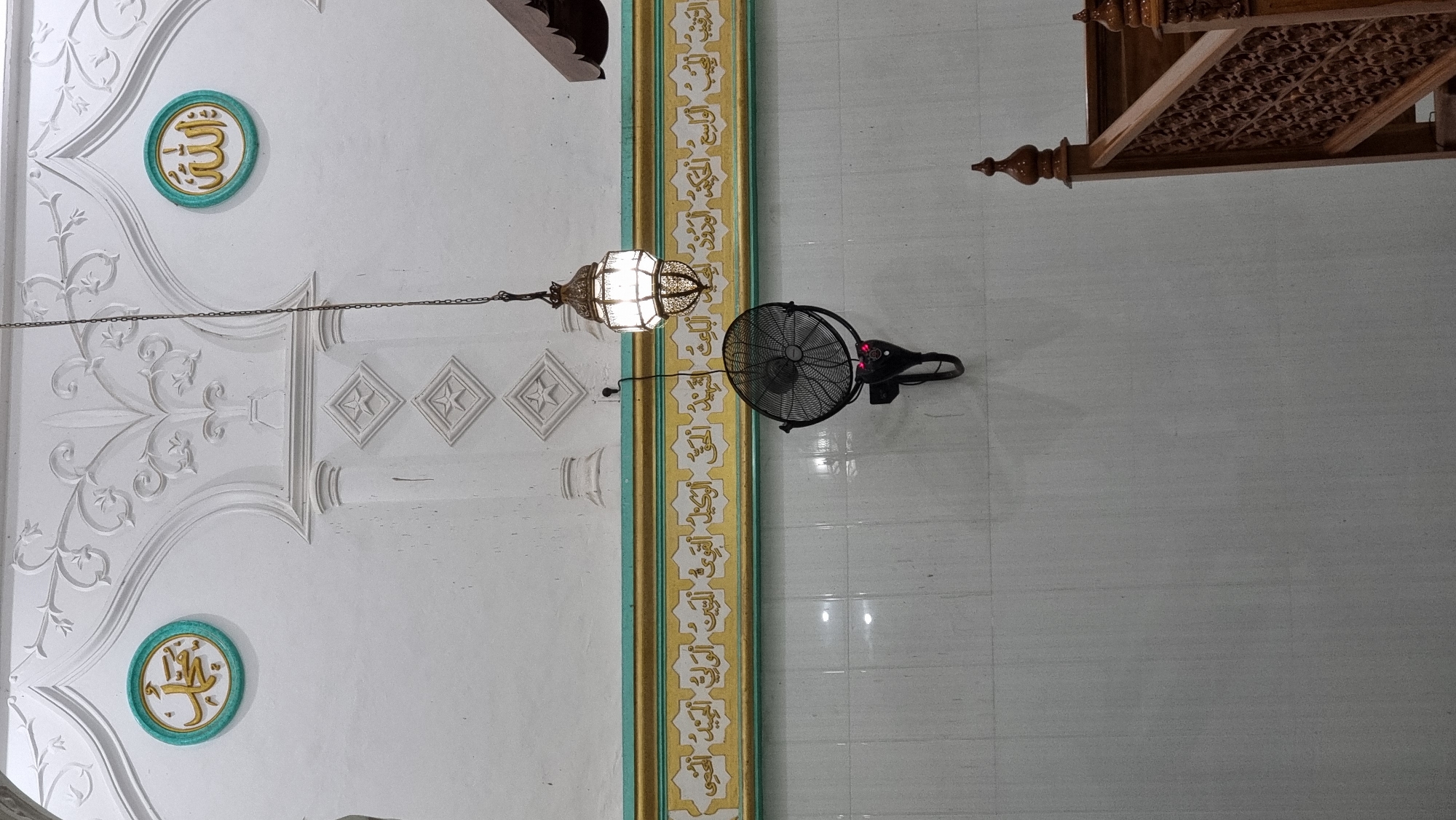 The image depicts the interior of a mosque, likely in Lapang Timu, Indonesia, given the context. The wall is white and adorned with intricate white plasterwork, featuring floral motifs and diamond-shaped insets. Two circular green and gold medallions with Arabic calligraphy are positioned high on the wall, one on the left with "Muhammad" and one on the right with "Allah." Below these medallions, a horizontal band of gold and green trim runs across the wall, above a section of tiled wall. A vintage lantern, illuminated, hangs from a chain in the center of the image, casting light. Below the lantern, a black oscillating fan is mounted on the wall. To the right, a portion of a wooden staircase with carved railings is visible, suggesting a minbar or elevated area. The overall impression is of a serene and traditionally decorated religious space.