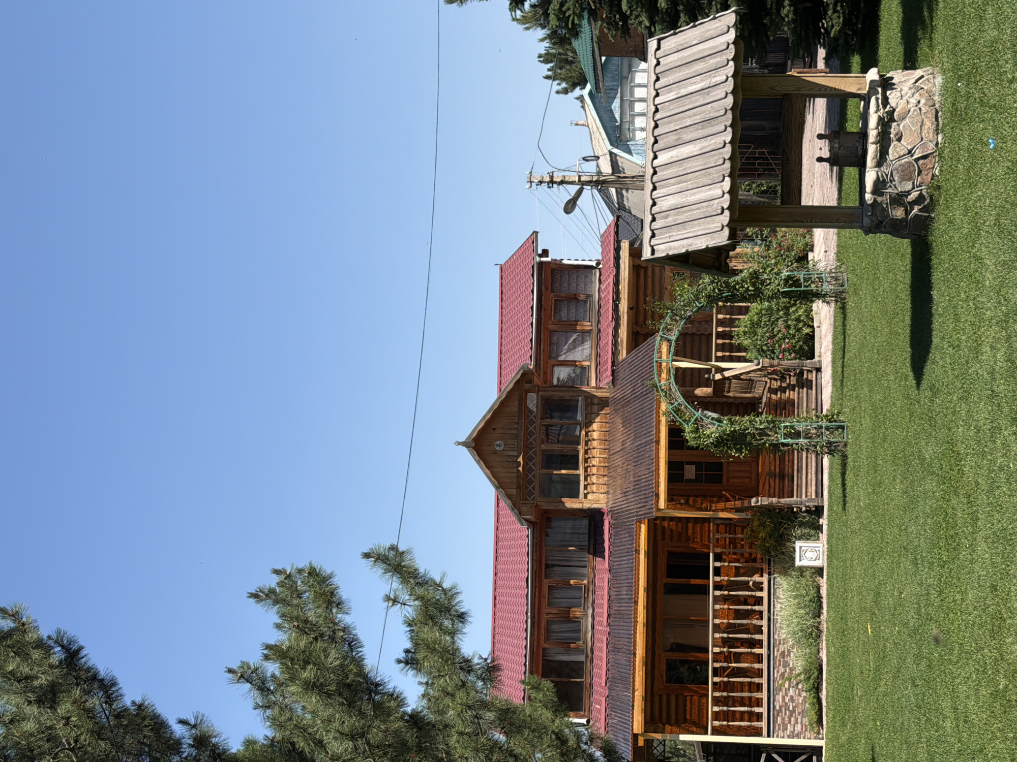 An outdoor, eye-level, full shot captures a sunny day scene with a wooden house and a well. The house is two stories tall with a red metal roof and log construction. A wooden porch with railings wraps around the front of the house. An arched trellis covered in greenery stands next to the front steps. To the right of the house is a stone and wood well with a bucket inside. A large pine tree is visible on the left side of the frame. The background shows other houses and trees under a clear blue sky. The scene is set in the city of Unknown, Unknown.