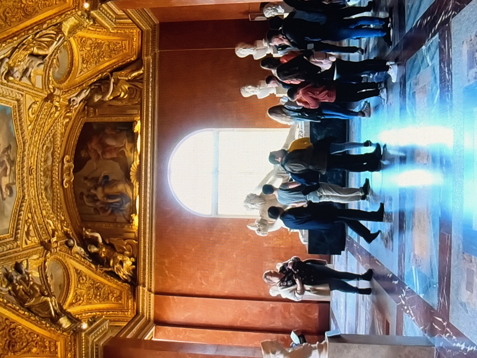 This is a medium shot of a group of people in a museum room. The room has ornate gold ceilings and red walls. There are several classical sculptures displayed throughout the room, including busts on pedestals. The floor is made of polished marble with a reflective surface, which is lit by natural light coming from a large arched window. The people are diverse in age and gender, and they appear to be looking at the sculptures. Some people are standing in groups, while others are alone. The overall atmosphere of the scene is one of quiet observation and appreciation of art.