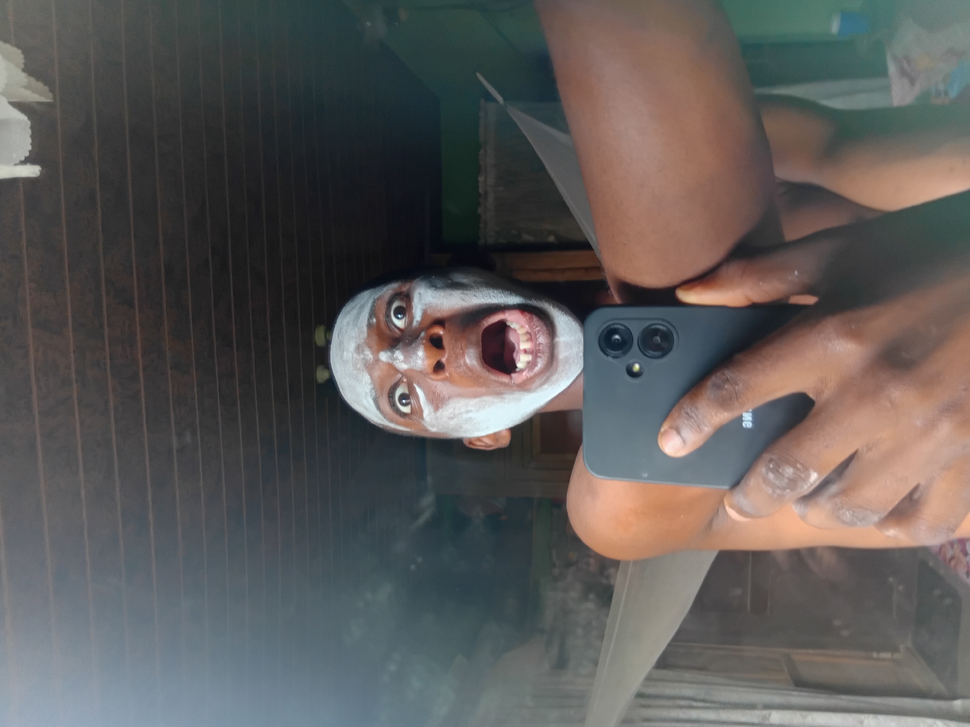 The image captures a close-up, selfie-style photograph of a young Black male with a surprised or shocked expression. He is applying a white substance, likely a facial mask, to his face, leaving his forehead, nose, and cheeks coated. His mouth is open, revealing his teeth, and his eyes are wide. He is holding a dark-colored smartphone in his right hand, pointed towards himself to take the picture. His left arm is visible, extended outward. The background suggests an indoor environment with a wooden slatted ceiling and a green wall. The scene appears to be in a residential setting in Nkpor, Nigeria, evidenced by the ambient light and common household features.