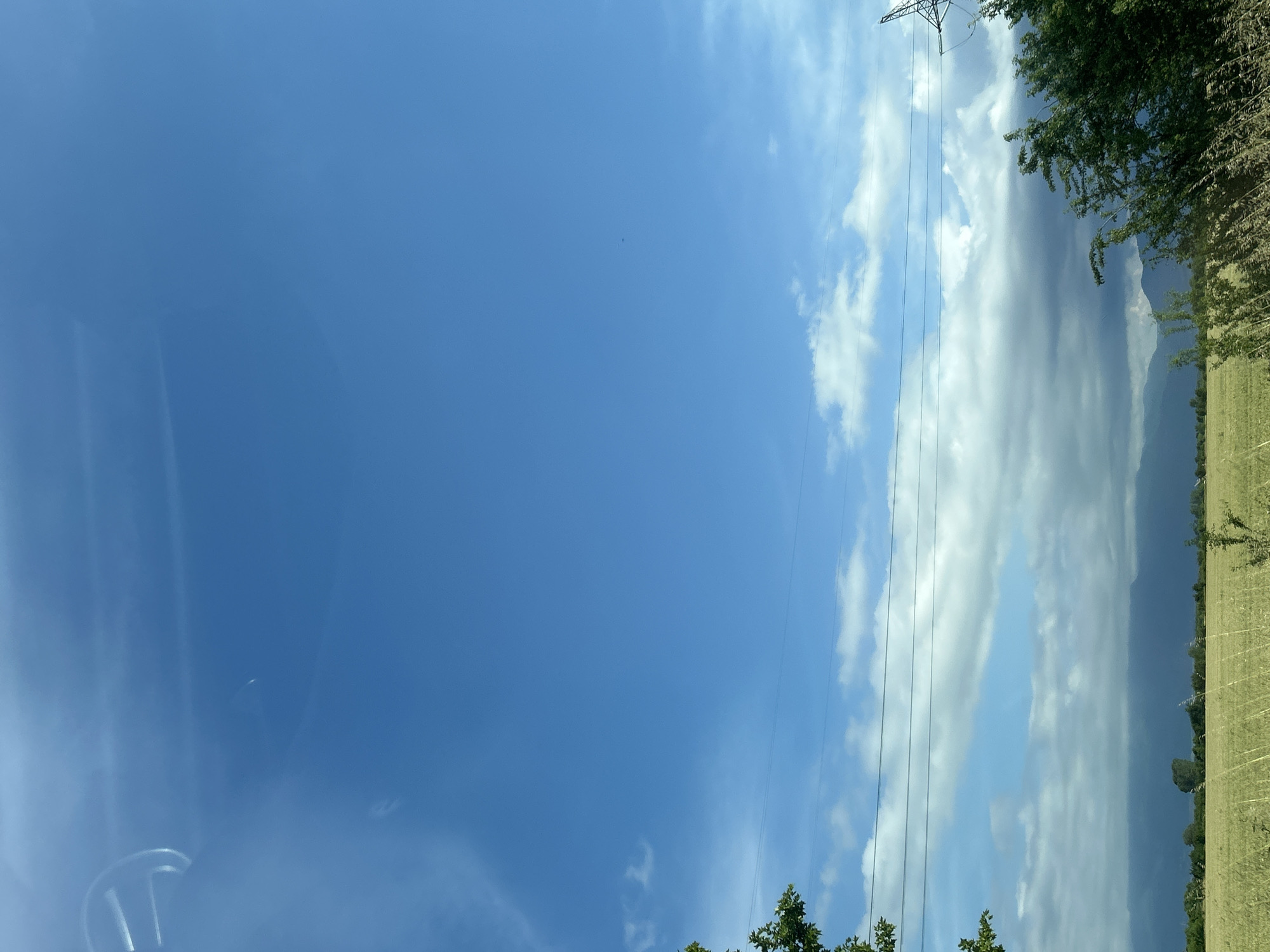 This landscape photograph, likely taken from a vehicle in Malo Konare, Bulgaria, presents a wide outdoor view under a bright daytime sky. The upper portion of the image is dominated by a vast expanse of clear blue sky. Towards the right side, scattered white cumulus clouds are visible, intermingling with some larger, darker grey cloud formations along the distant horizon.
A prominent electricity pylon stands on the far right, with multiple thin power lines extending horizontally across the mid-sky. In the mid-ground, a range of large mountains emerges, their peaks partially veiled by a slight haze or distant atmospheric conditions. Below the mountains and extending across the lower right of the frame is a field of dried grass or crops, indicative of agricultural land. Green trees with dense foliage line the edge of this field and also appear in the bottom left foreground, framing the scene.
The photograph captures a sunny day, with ample natural light illuminating the environment. The weather appears fair, characterized by a mix of bright sky and varying cloud cover. A subtle reflection or part of a car's interior, possibly a steering wheel, is visible in the bottom left corner, suggesting the image was captured from inside a vehicle. There are no discernible human figures or visible text in the scene, conveying a sense of serene, natural countryside in a rural Bulgarian setting.