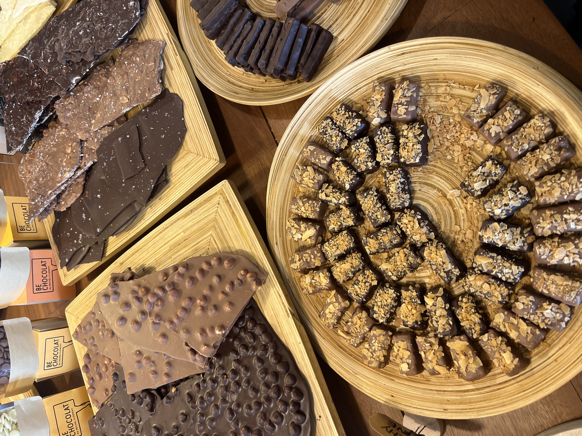 This high-angle shot displays a variety of chocolates arranged on wooden trays, likely in a shop or market setting. The overall scene is indoors, with warm lighting suggesting a cozy atmosphere.
On the left, three rectangular wooden trays are filled with different types of chocolate bark. The top left tray holds dark chocolate bark with what appears to be crushed nuts or sea salt. Below that, another tray features milk chocolate bark also adorned with visible inclusions. The bottom left tray showcases large pieces of milk chocolate bark with whole hazelnuts, and next to it, dark chocolate bark with a similar hazelnut inclusion.
To the right, a large circular wooden tray is generously filled with small, rectangular chocolate pieces. These chocolates are mostly dark or milk chocolate, each topped with a generous amount of golden, crunchy pieces, possibly crushed biscuits, nuts, or toffee bits. These pieces are arranged in a circular pattern around the tray.
In the background, another small circular wooden tray contains neatly stacked, elongated dark chocolate sticks or fingers.
Several small, light brown cardboard boxes are visible, partially obscured, beneath the chocolate bark trays on the left. The visible text on these boxes reads: "BE CHOCOLAT by MICHEL CLEMENT." These boxes suggest branded packaging for the chocolates. The textures of the chocolates range from smooth to rough with various toppings, creating an appetizing display of artisanal confections.
