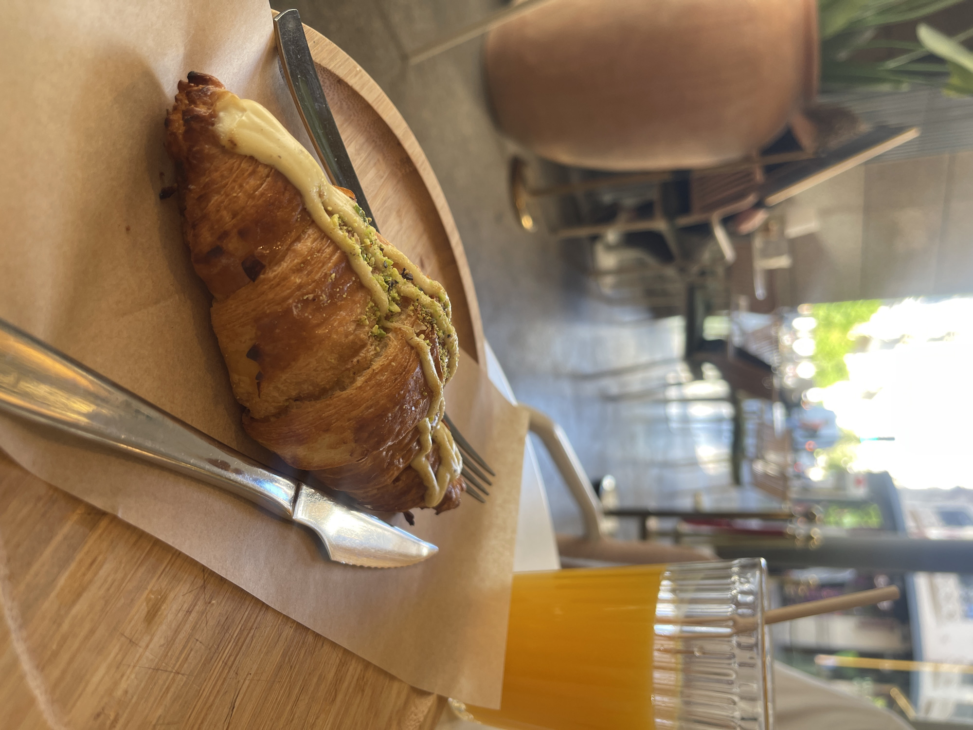 On a wooden table, in a bright, possibly outdoor seating area in València, Spain, sits a croissant, drizzled with a white sauce and green pistachio crumbles, on a piece of brown parchment paper, along with a knife and fork. Next to the croissant, slightly out of focus, is a glass of orange juice with a straw. In the background, blurred but discernible, are tables, chairs, and people, suggesting a cafe or restaurant setting. The exterior hints at urban landscaping with trees and buildings visible through the seating area. A large, unglazed earthenware pot with foliage is visible behind the croissant.