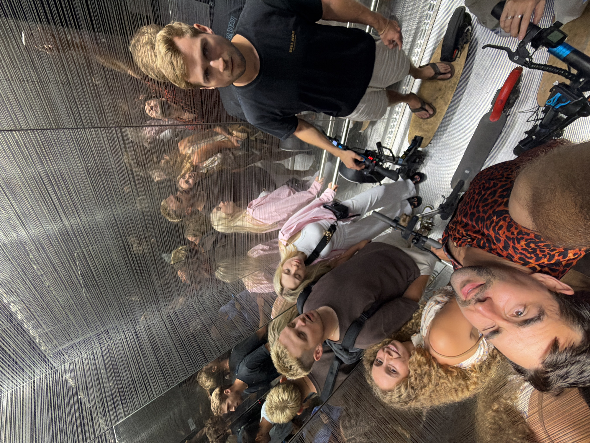The image depicts a group of young adults inside what appears to be an elevator with mirrored walls and a textured, metallic ceiling. Multiple reflections of people and objects are visible, creating a disorienting effect. In the foreground, a man with short blond hair wearing a black t-shirt and light-colored shorts stands on an electric scooter. His right hand is on the handlebar of the scooter, and he is looking directly at the camera. Behind him, other individuals are reflected in the mirrors, some appearing to be on scooters as well. A woman with long blonde hair is visible in a pink top and white pants. Another woman with curly brown hair is smiling at the camera. The overall scene suggests a casual gathering of friends, possibly exploring the city of Tel-Aviv, Israel, on electric scooters. The interior of the elevator seems modern and sleek, with the metallic texture of the walls and ceiling being a prominent feature.