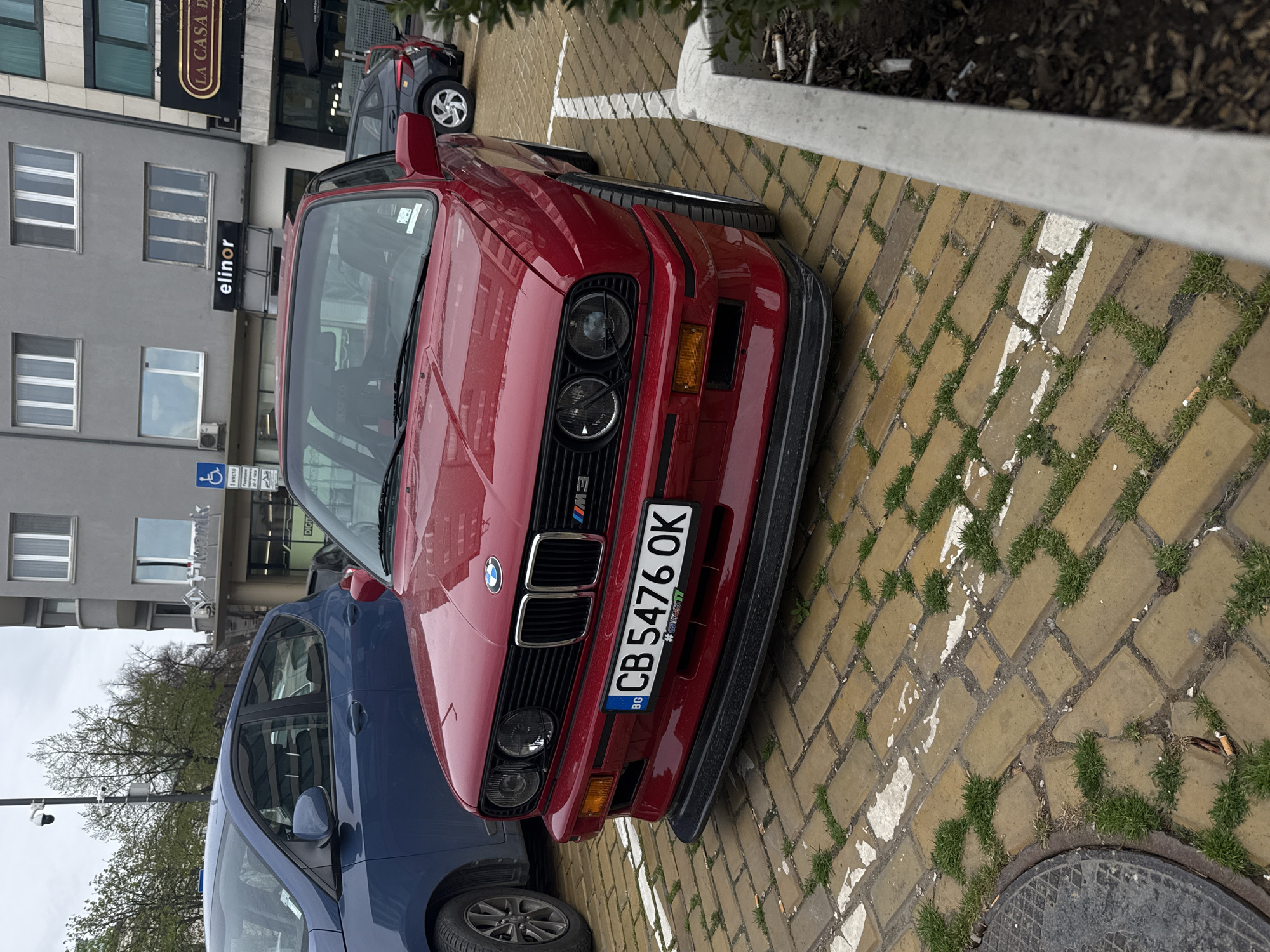 This full-shot image captures a vibrant red, classic BMW M3 (E30 generation) parked outdoors on a paved surface, viewed from a slightly elevated angle. The car is positioned at an angle, with its front grille and license plate prominently displayed. The parking surface consists of light brown paving stones with green grass growing in the gaps, and white lines indicating parking spaces, which are somewhat worn.
In the immediate foreground, a part of a blue car is visible to the left, and in the background, another dark grey car is parked. The setting appears to be an urban area, with multi-story buildings visible behind the parked cars. One building is grey with multiple windows, and another, to its right, features a dark facade with outdoor seating, possibly for a cafe or restaurant.
Notable details include:
- The red BMW M3 features a prominent "M3" badge on its grille and a black front spoiler. Its headlights are circular, and the turn signals are amber.
- The license plate reads "BG CB 5476 OK" and has a small sticker below it that says "#e30mania".
- On the building in the background, a partial sign reads "LA CASA D" and another sign clearly displays "elinor". A disabled parking sign with a wheelchair pictogram is also visible.
- Through the windshield of the red BMW, a "sparco" branding can be seen, likely on a seat or harness.
- The time of day is daytime, with soft, diffused lighting suggesting an overcast sky.
The overall scene depicts a classic car parked in a bustling city environment, possibly in Sofia, Bulgaria, given the license plate and provided context.