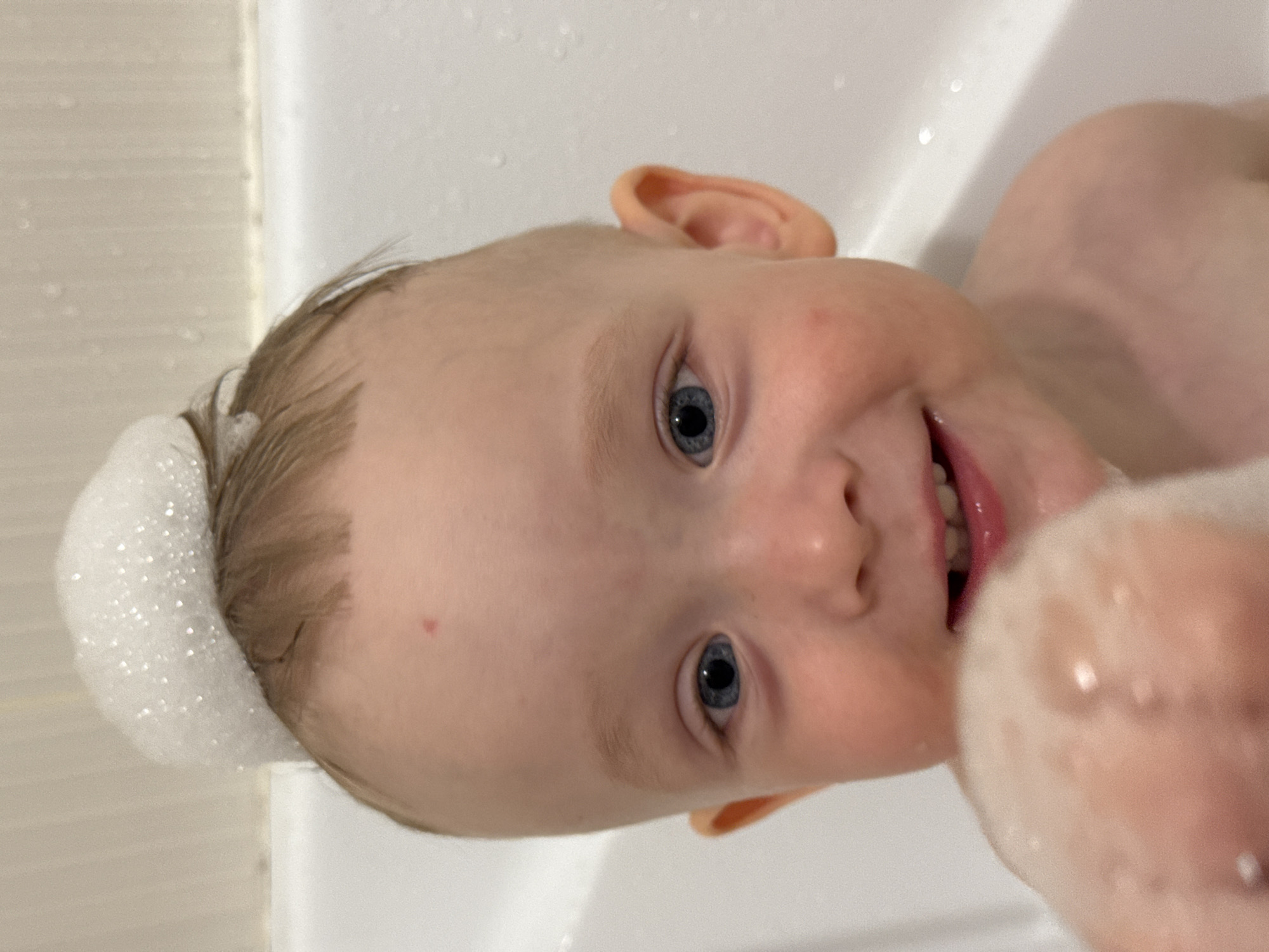 A close-up, waist-up, indoor shot shows a fair-skinned toddler with blue eyes and light brown hair in a bathtub. A small amount of white bubble bath foam is perched on top of the toddler's head, and a larger clump of foam is being held in front of the toddler's face, partially obscuring it. The toddler is smiling, revealing small teeth. The background is a white bathtub with some water droplets visible on the surface. The toddler appears to be in a private residence. The image does not provide information about the location in Chaikovsky, Russia.