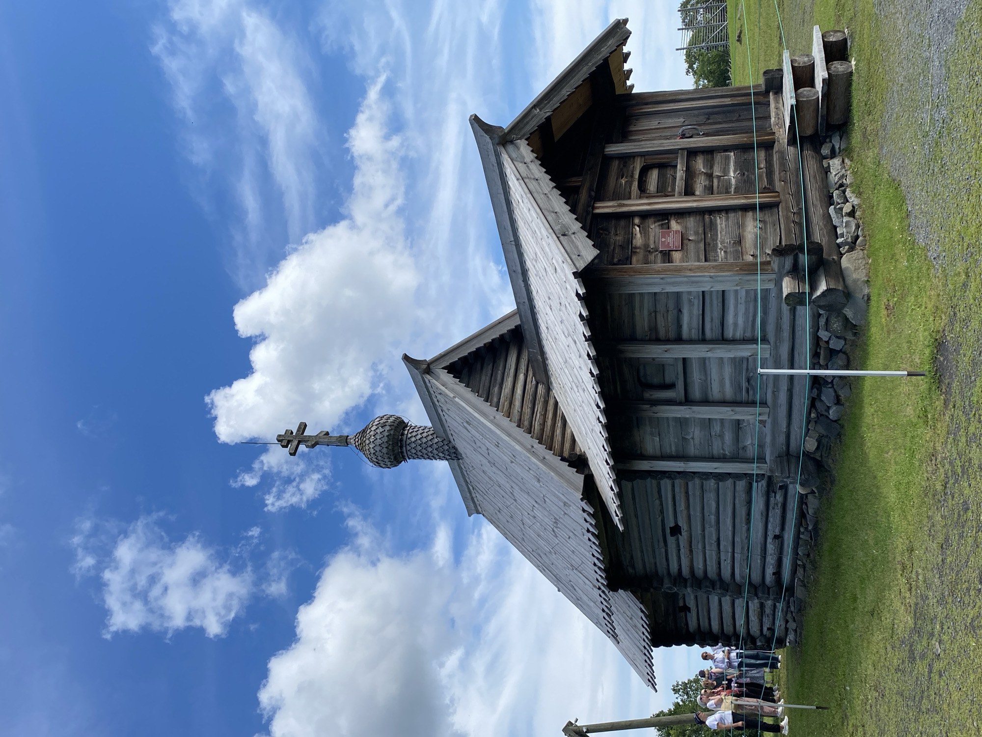 A wooden church stands in the foreground against a bright blue sky with scattered white clouds. The church is constructed from logs and features a multi-tiered roof, with a central dome topped by a cross. The structure has a rustic appearance, with visible wood grain and weathered timber. A small group of people, dressed in casual clothing, are standing to the left of the church, observing it. The ground around the church is covered in green grass, and a low rope barrier is visible in front of the building. The background shows a wooden fence and trees in the distance, suggesting an outdoor, possibly historical, setting. The location is Boyarshchina, Russia.