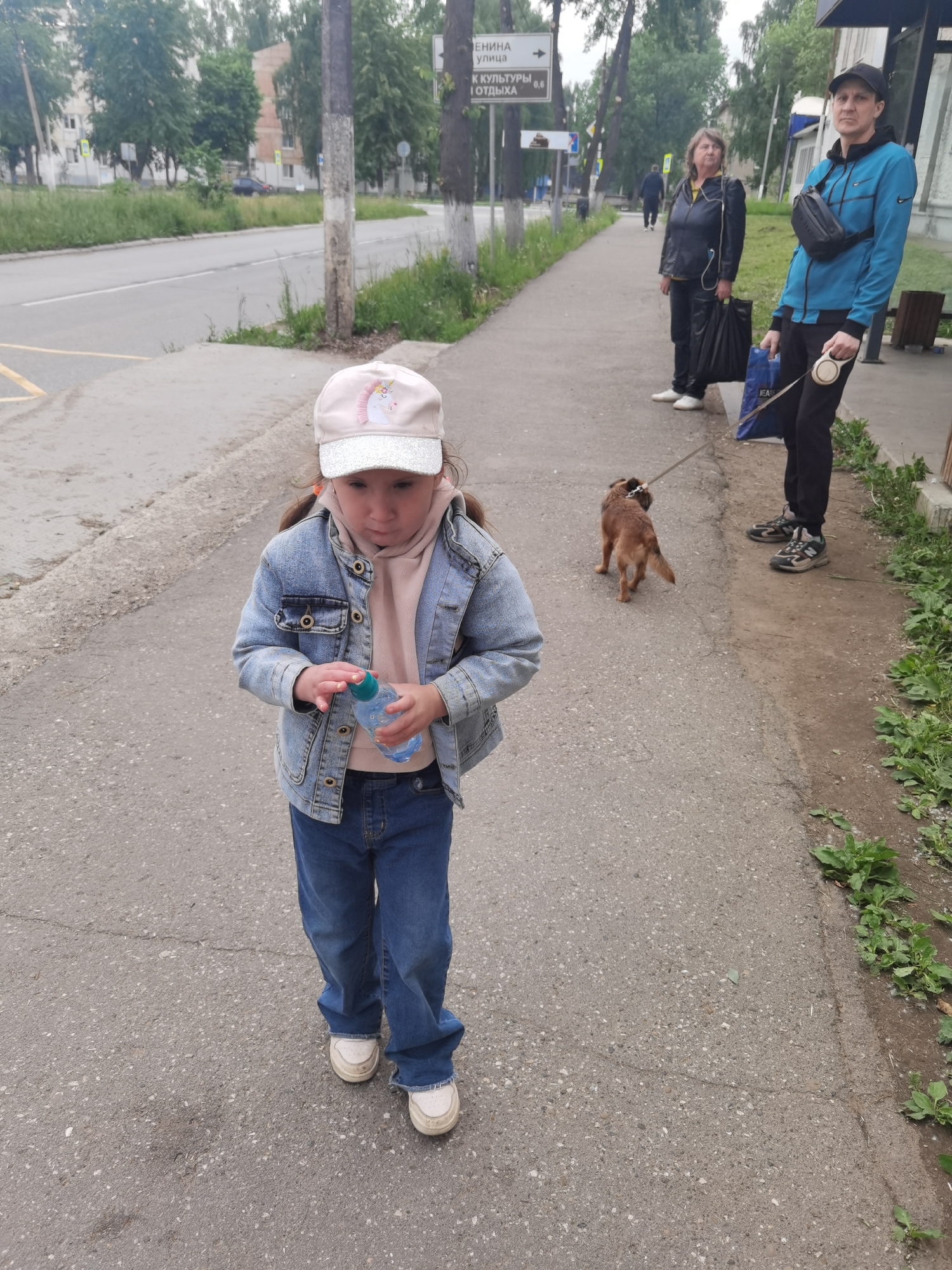 The full-shot outdoor photograph captures a street scene in Chaikovsky, Russia, featuring a young girl in the foreground, a small dog, and two adults in the background.
In the center foreground, a young girl walks on a grey asphalt sidewalk. She wears a light blue denim jacket over a pink hoodie, blue jeans, and white sneakers. On her head is a light pink baseball cap with a white brim and a unicorn emblem. She holds a small plastic water bottle with a blue cap in both hands, looking down at it. Her expression appears focused or slightly serious.
Behind her, to the right, a small, brown, short-haired dog walks on a retractable leash held by a man. The man, on the far right, wears a blue jacket with a Nike logo, black pants, a black baseball cap, and athletic shoes. A black fanny pack or chest bag is strapped across his front. He holds the dog's leash in one hand and a blue "JEAN" branded shopping bag in the other. To his left, an older woman stands, wearing a black jacket, dark pants, and white sneakers, holding a black shopping bag. Both adults look forward with neutral expressions.
The setting is an urban or suburban area. To the left of the sidewalk is a paved road with some yellow markings. Further in the background, residential buildings are visible amidst numerous green trees, suggesting a cloudy or overcast day. Tall utility poles line the sidewalk. A street sign in Russian is prominent on one of the poles, pointing towards "ЛЕНИНА УЛИЦА" (Lenin Street) and indicating "К КУЛЬТУРЫ И ОТДЫХА" (To Culture and Recreation) at a distance of "0.6" (likely kilometers). The ground to the right of the sidewalk is unkempt, with patches of grass and weeds. The overall atmosphere is mundane, capturing a moment of everyday life.