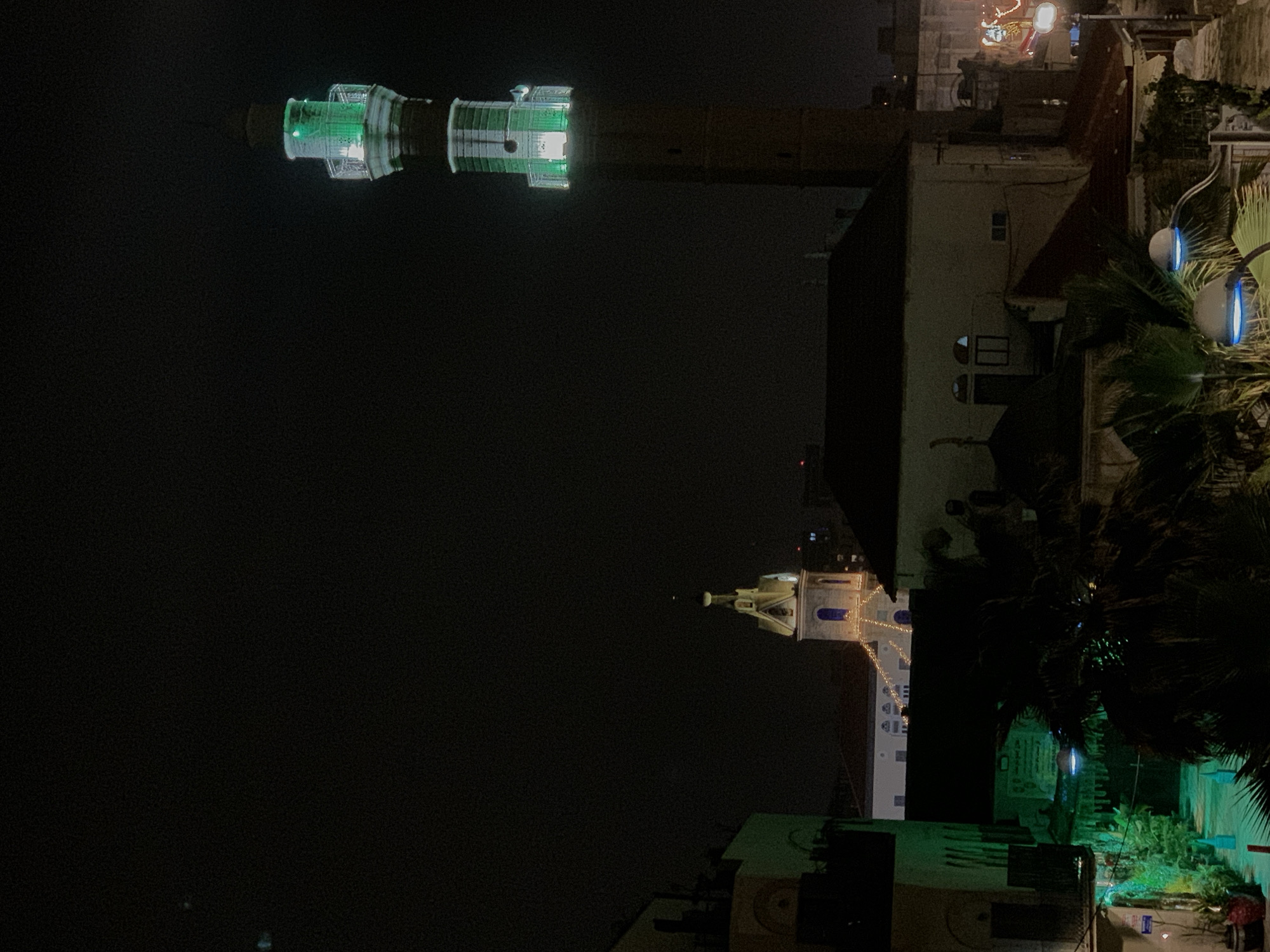 The image captures a night scene in Tel-Aviv, Israel, showcasing two prominent structures. On the left is the lit-up minaret of the Hassan Bek Mosque, decorated with bright green lights. Another building is visible, with a tower adorned with white and blue lights, possibly an adjacent church. The surrounding environment includes residential buildings and palm trees, which is lit by street lights. The sky is dark.