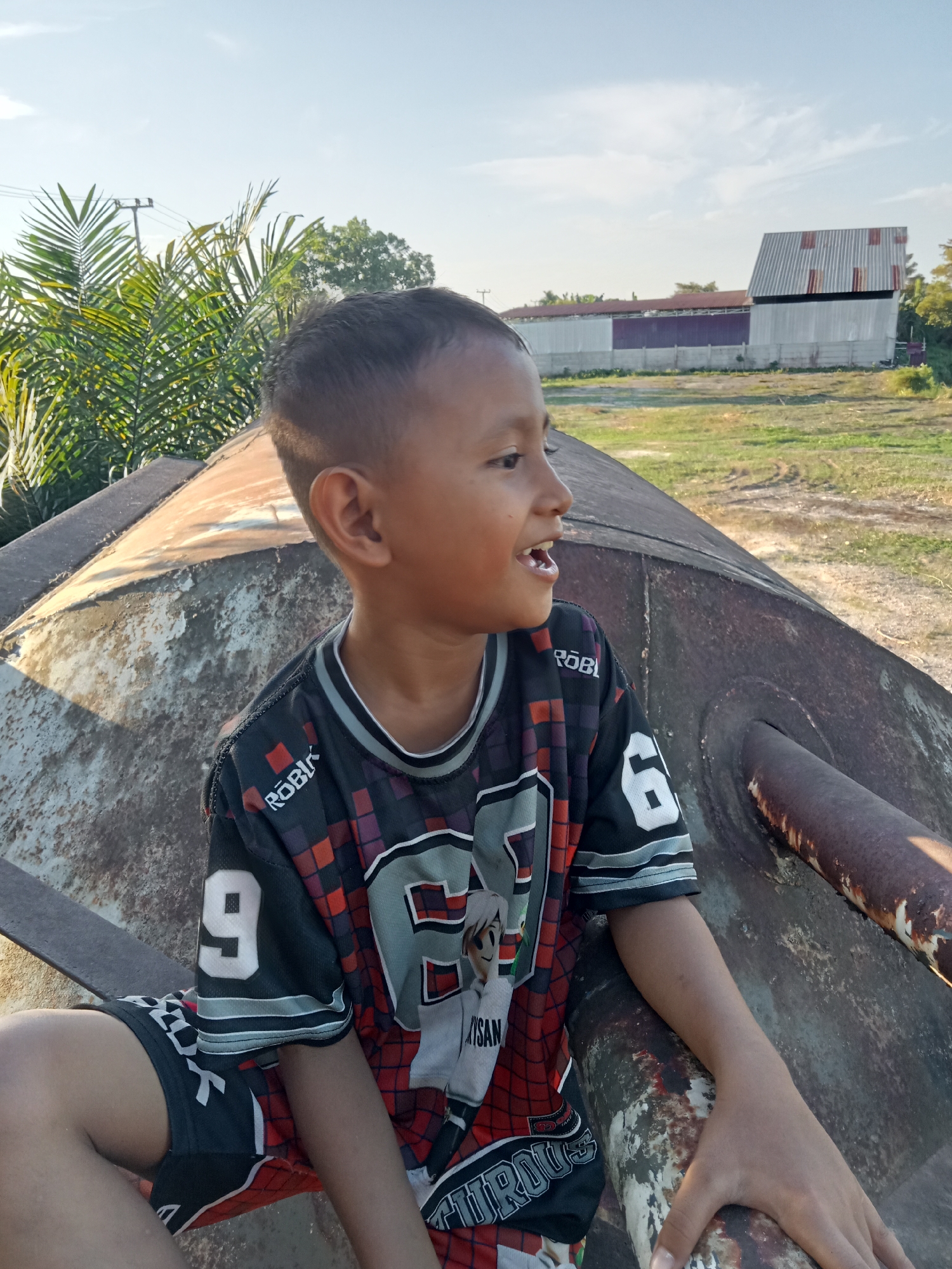 A young boy with short dark hair, wearing a black and red jersey with white numbers 9 and 6, and dark shorts, sits on a rusty metal structure outdoors during the day. He is looking to his right with his mouth open. In the background, palm trees, a building with a corrugated metal roof, and a grassy field are visible under a clear blue sky. The overall scene suggests an outdoor setting in Kualu, Indonesia.