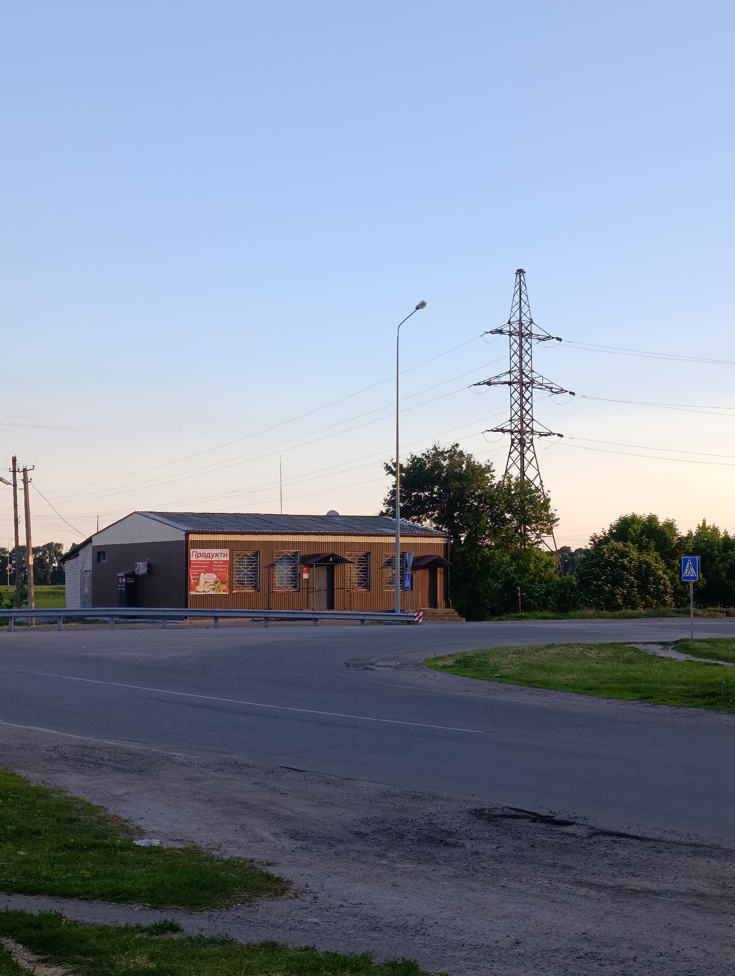 The image captures a static, outdoor scene at what appears to be an intersection or curve on a road, likely in a rural or semi-urban area of Krasnohorivka, Ukraine. The time of day is late afternoon or early evening, characterized by a clear, light blue sky that subtly transitions to a warmer, peachy hue near the horizon, and soft, warm light illuminating the scene from the right.
The primary subject is a single-story building on the left side of the road, which appears to be a shop. The building has a brown corrugated facade and a grey roof, with several windows protected by decorative metal grilles. An air conditioning unit is visible on its left side. A large red and white banner is prominently displayed on the front of the building, featuring the Cyrillic word "ПРОДУКТИ" (PRODUKTY), which means "Groceries" or "Products." Below this, smaller text and images of various food items like milk, bread, and cheese are visible, indicating the nature of the store. A trash bin is placed beside the building's left corner.
In front of the building, a metal guardrail separates it from the asphalt road, which curves to the right. The roadside and median areas are covered with green grass and dirt. A tall streetlight pole stands between the building and a massive, latticed electricity pylon, which dominates the right side of the sky. Several utility poles with wires are also present in the background. Dense green trees and bushes are scattered behind the building and around the base of the electricity pylon, adding natural elements to the setting. On the right side of the road, a blue square sign with a white triangle and a black symbol of a person walking indicates a pedestrian crossing. No people or animals are visible, and no specific activity is taking place; the scene is quiet and observational.