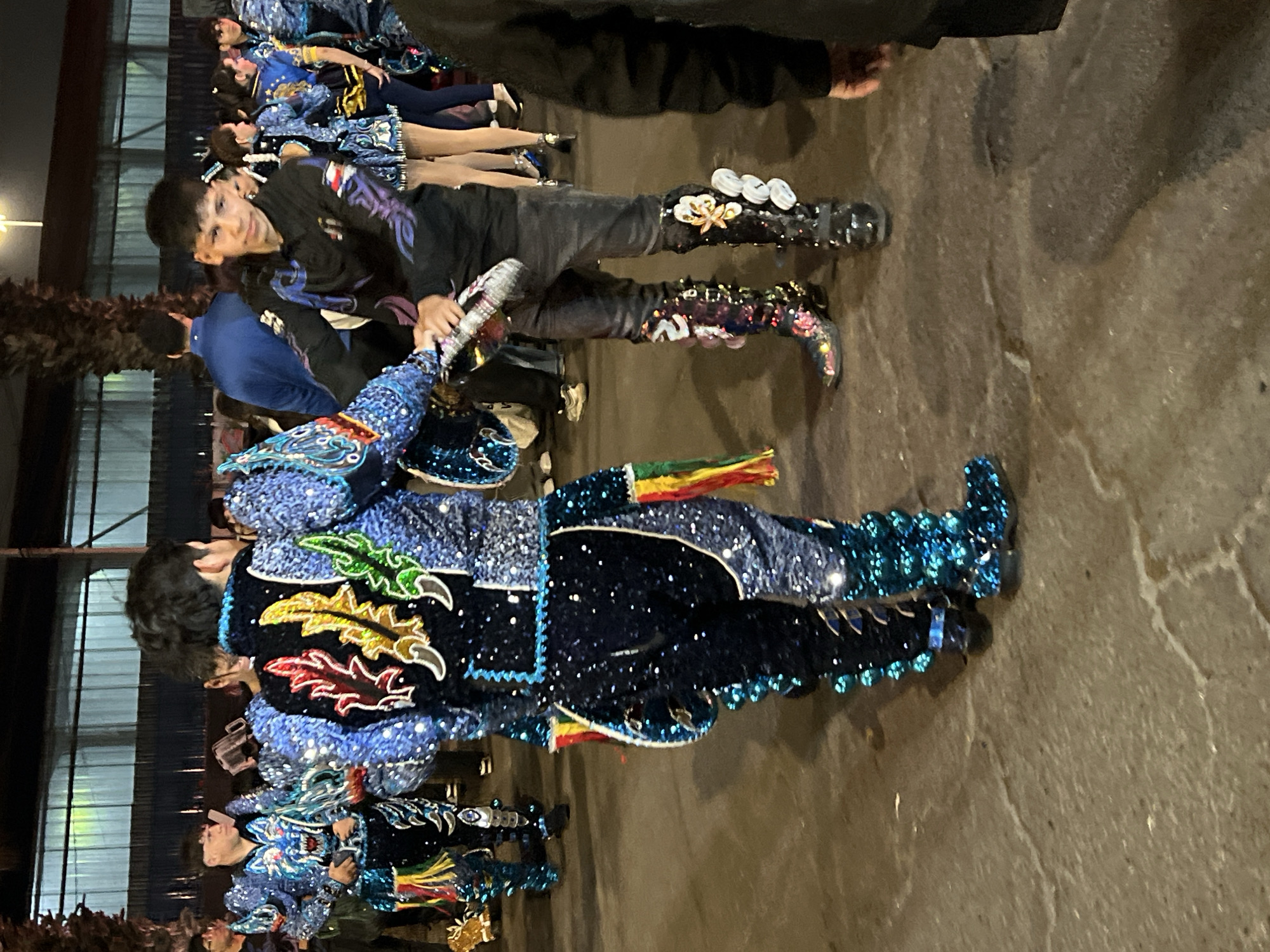 The image depicts a group of people in elaborate, sequined costumes standing on a paved ground, likely outdoors at night. The costumes are richly decorated with intricate patterns and brightly colored embellishments, suggesting a festive or ceremonial occasion, possibly a parade or performance. In the background, a large structure with horizontal ribbed panels and lighting can be observed, possibly a warehouse or a large tent. The lighting is artificial and casts shadows on the ground, indicating nighttime. One individual in the foreground is wearing a black long-sleeved shirt with purple and blue designs, dark jeans, and distinctive, ornate boots. Another person is wearing a blue, sequined costume with colorful flame-like appliques on the back. The overall atmosphere suggests a cultural event in the city of Arica, Chile, given the mention of the location.