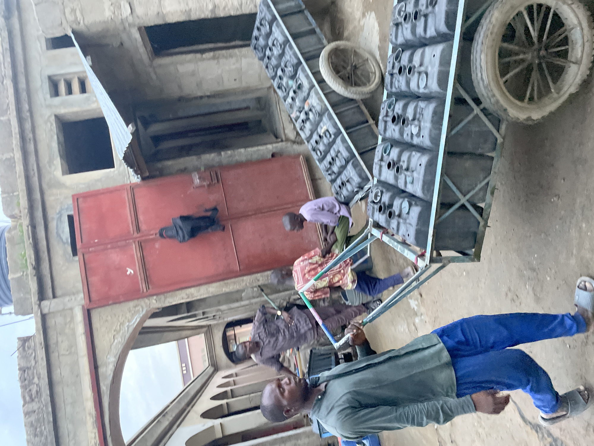 Three men are pushing a metal-framed cart filled with gray rectangular containers. The cart also has two spare tires attached to it. The men are dressed in casual clothing and sandals. The setting appears to be a street with older buildings on either side. A red metal gate is visible in the background. The location is Unknown, Unknown.