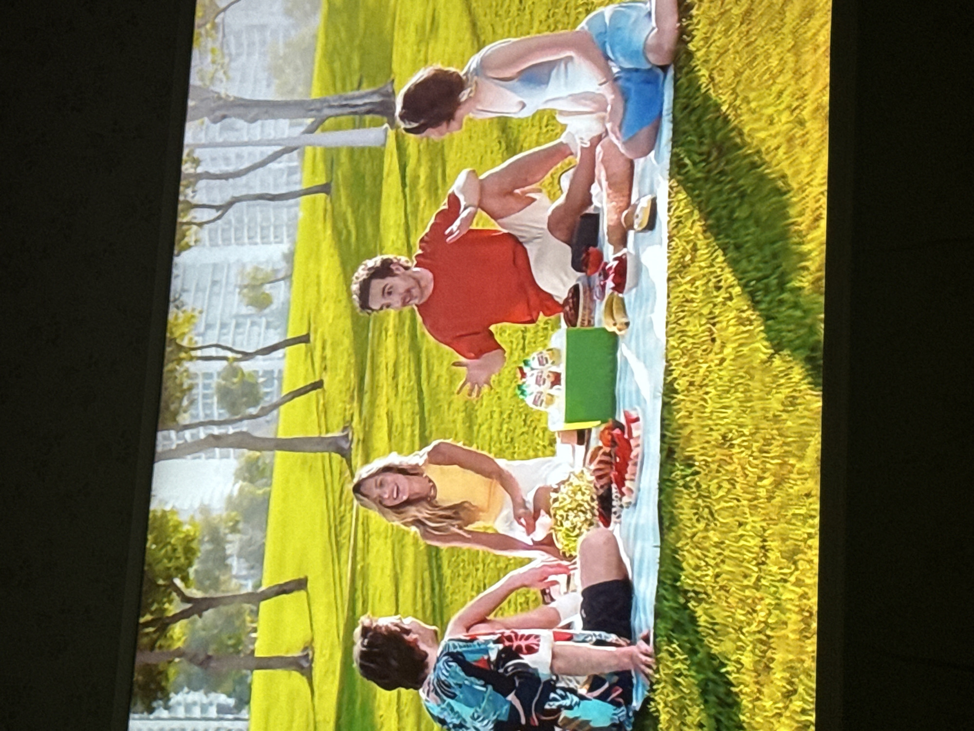 A group of four people are having a picnic in a grassy area with trees in the background. Behind the trees, there are buildings. The person closest to the bottom of the image is wearing a colorful floral shirt and dark shorts. Next to them is a person with blonde hair wearing a yellow shirt and white shorts. Across from them is a person in an orange shirt and white shorts. Another person is sitting next to them wearing a white tank top and blue shorts. There is a light blue picnic blanket spread out with food and drinks on it. There is a green box on the blanket as well. There is no indication this picture was taken in Mytishchi, Russia.