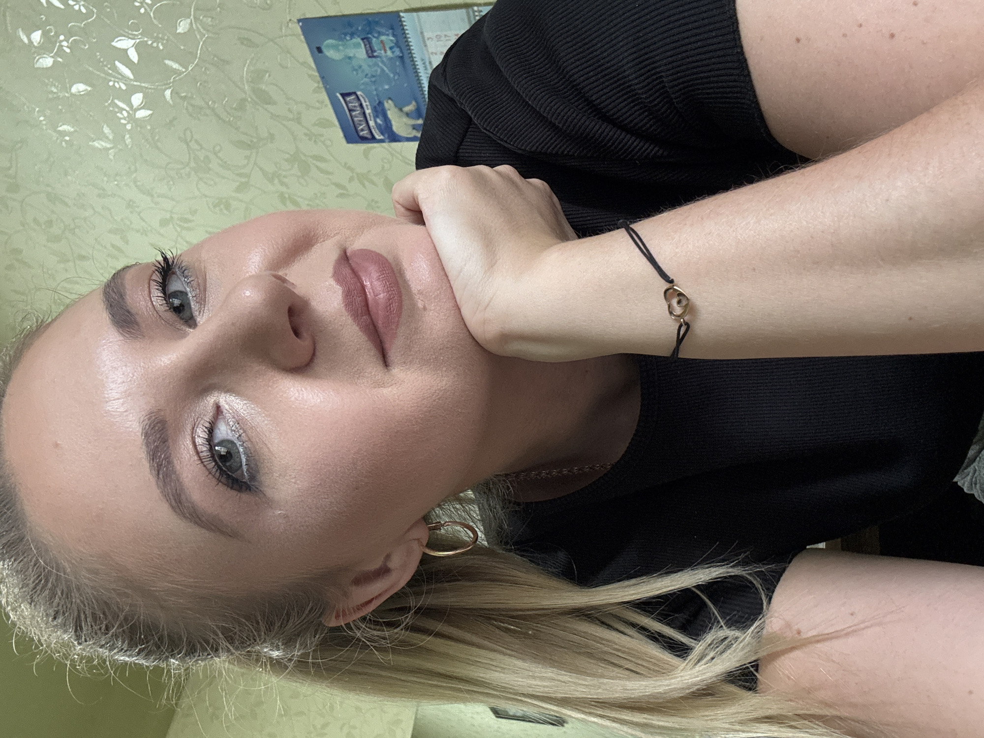 A blonde woman with her hair in a ponytail poses for a selfie indoors. She is wearing a black, ribbed t-shirt and a bracelet with a gold heart charm. Her hand rests on her chin as she looks at the camera. Behind her, a wall is covered in a green floral-patterned wallpaper, and a calendar featuring polar bears and water bottles is hanging on the wall.